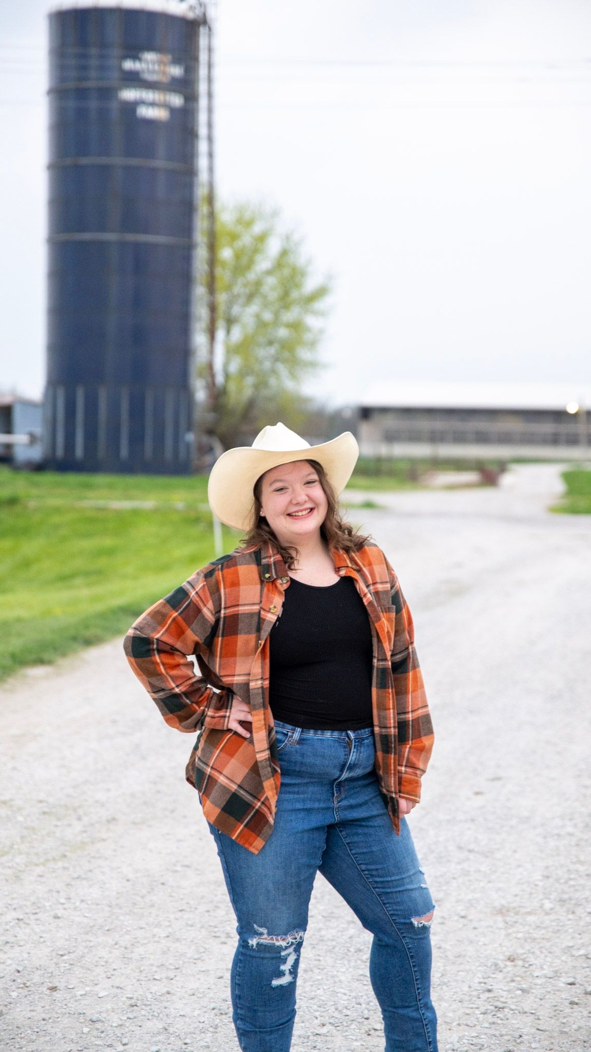 Person in plaid shirt and cowboy hat poses on gravel road with grain silos in background.