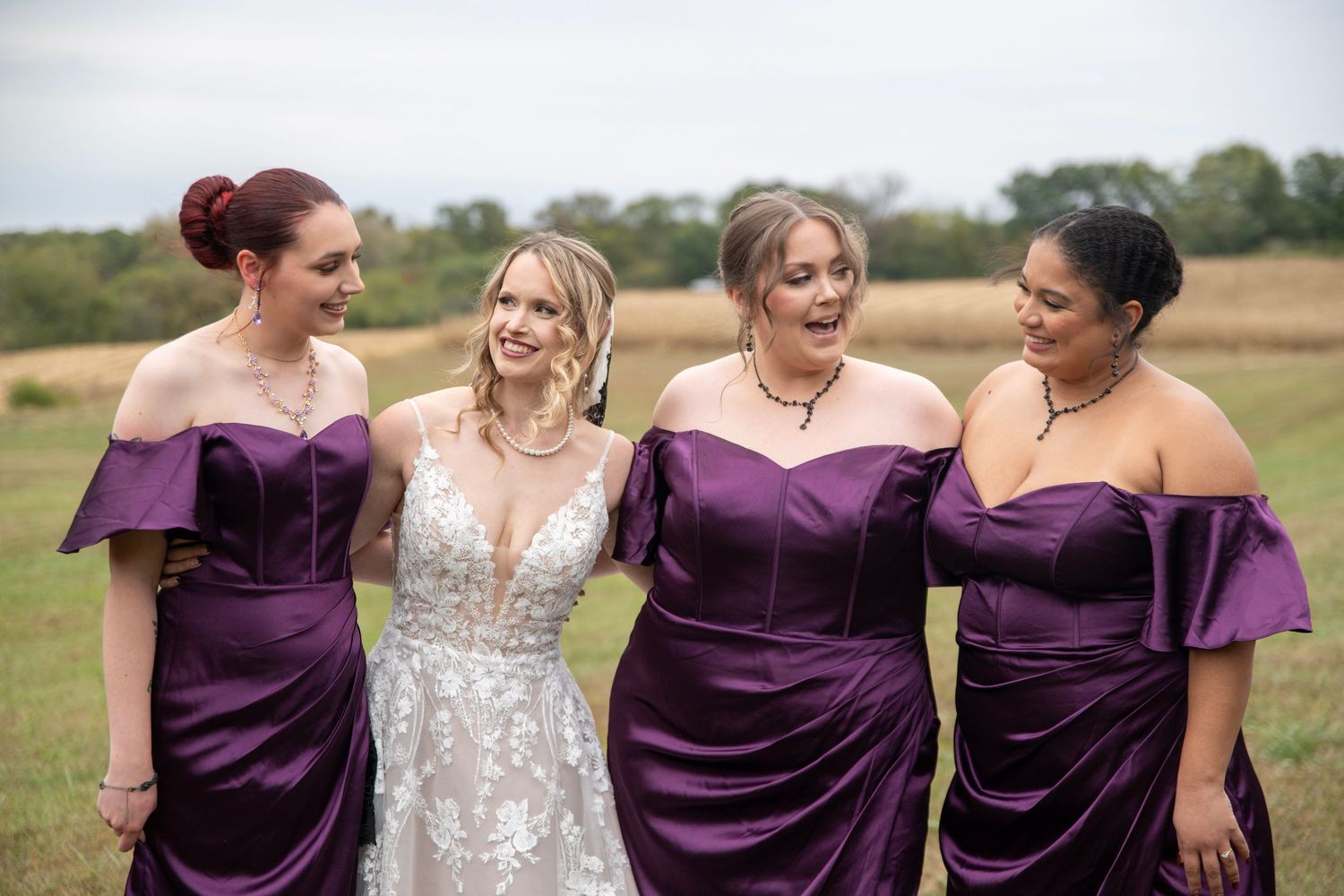 Bride in lace gown poses with bridesmaids wearing elegant purple off-shoulder dresses in an outdoor field setting.