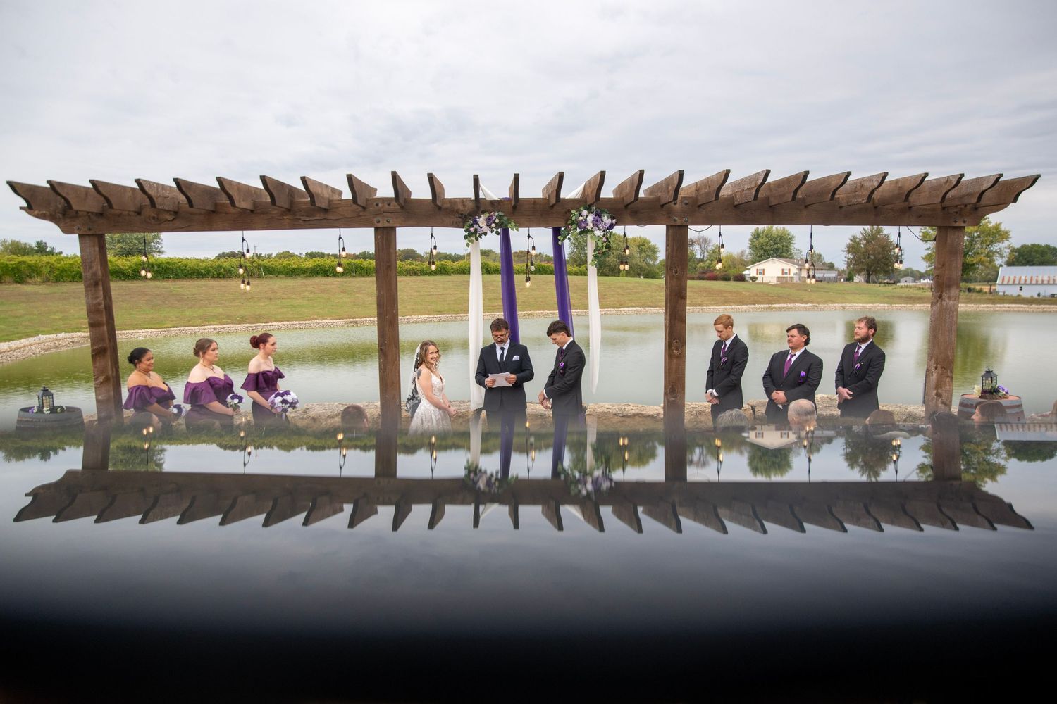 Reflection shot of wedding party lined up under wooden pergola by water.