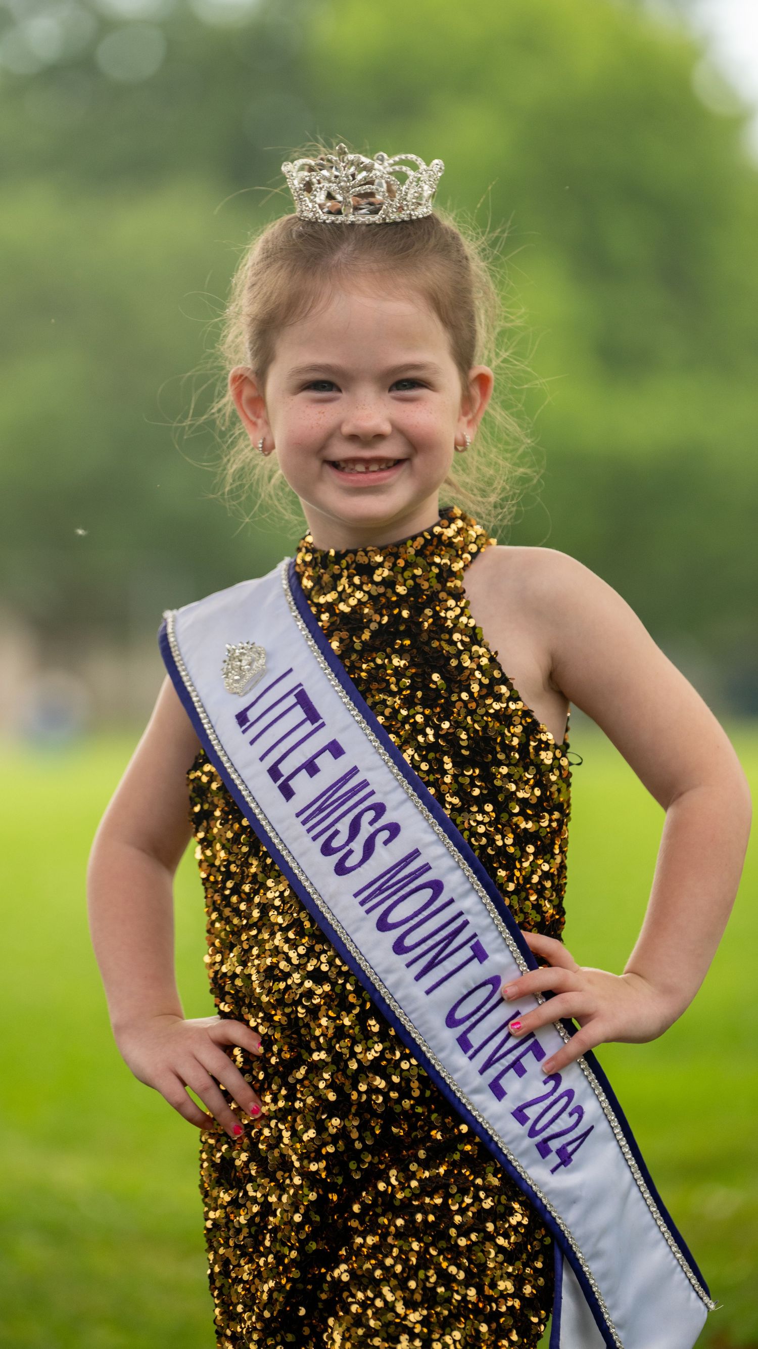 Young pageant contestant wearing a sparkly black and gold dress with sash outdoors.