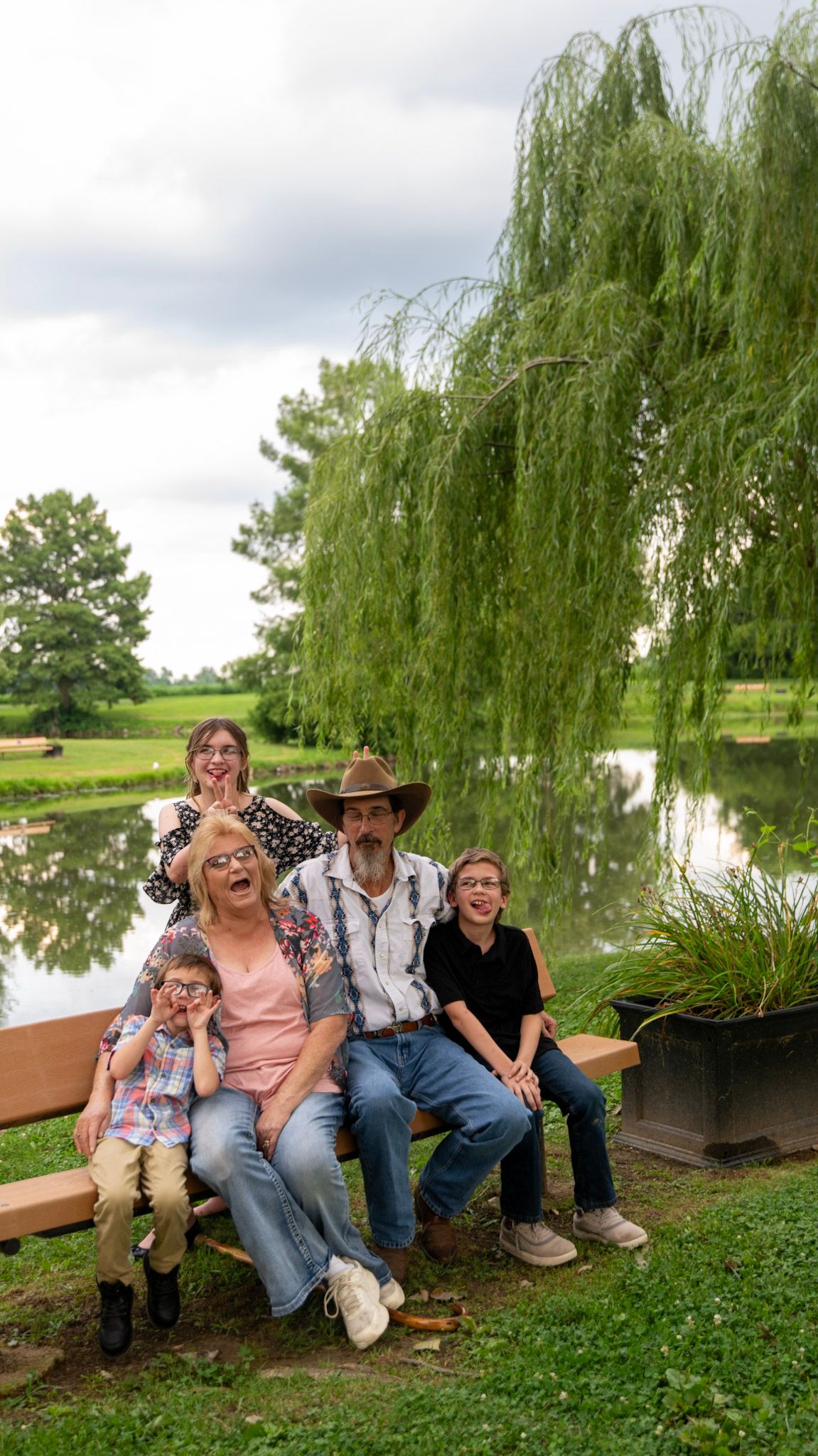 A family sits together on a wooden bench by a pond with a weeping willow tree in the background.