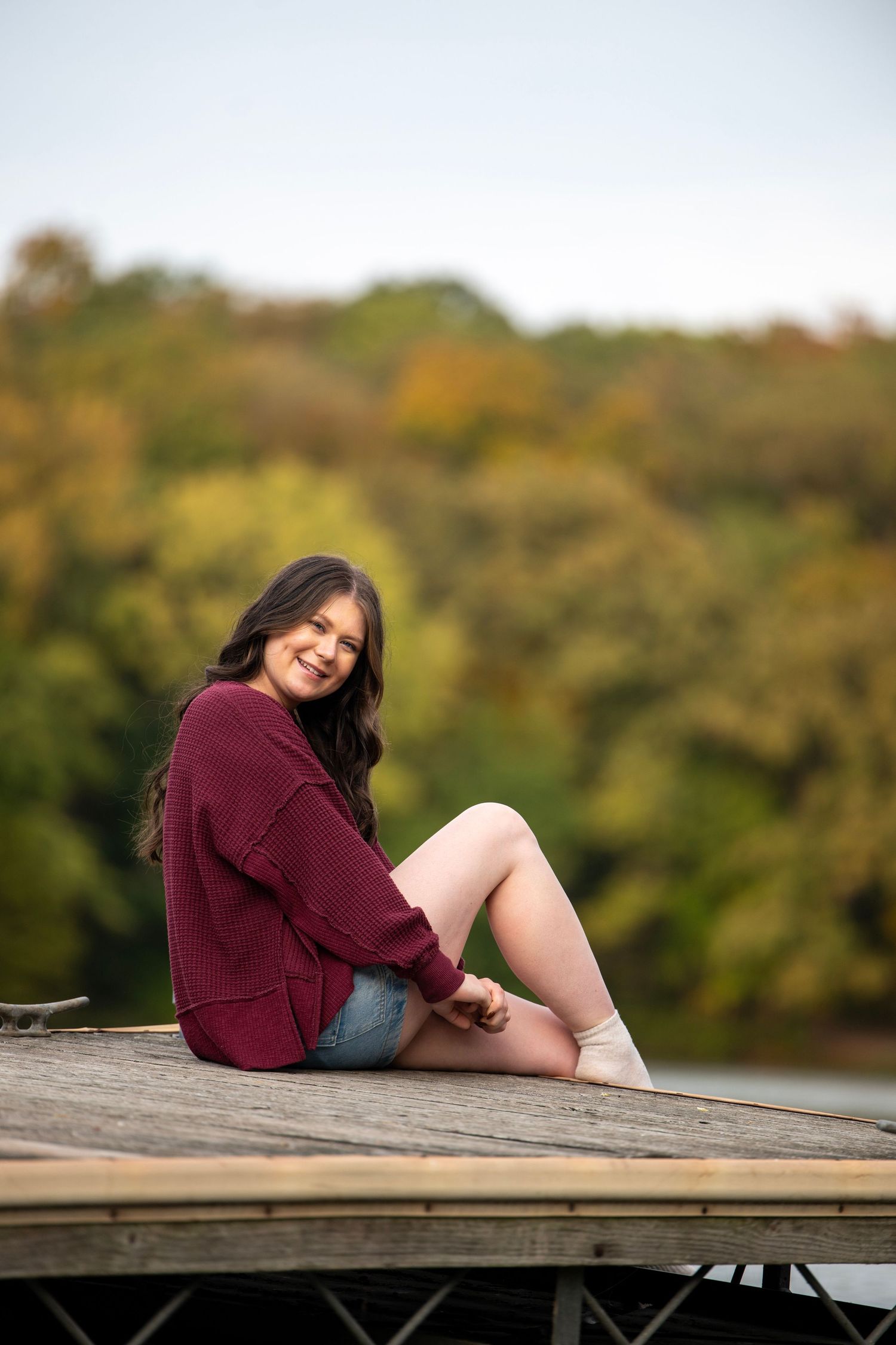 A person in a burgundy sweater lounges on a wooden dock surrounded by autumn foliage.