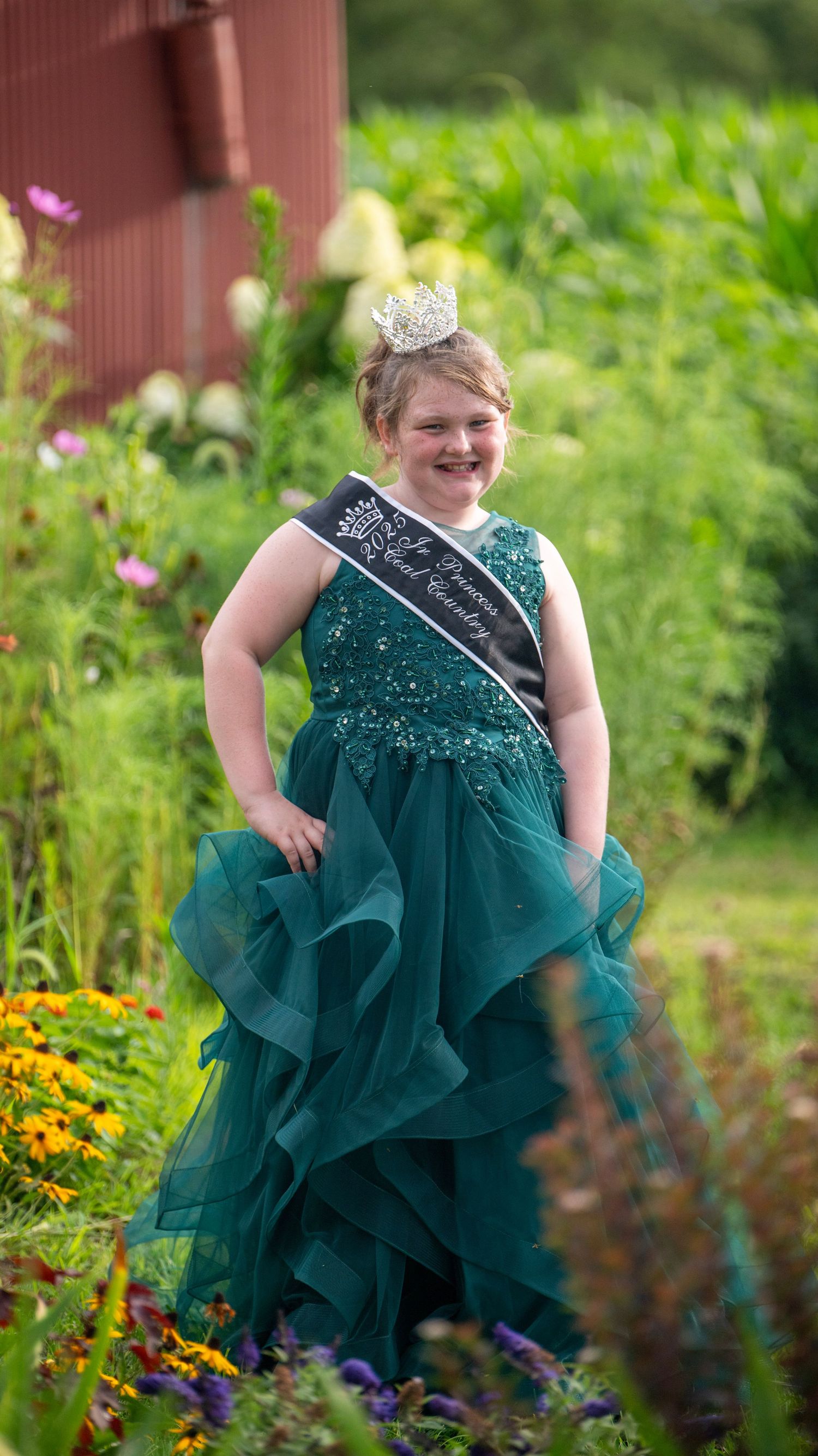 Person in an emerald green formal gown poses in a lush garden setting with yellow flowers.