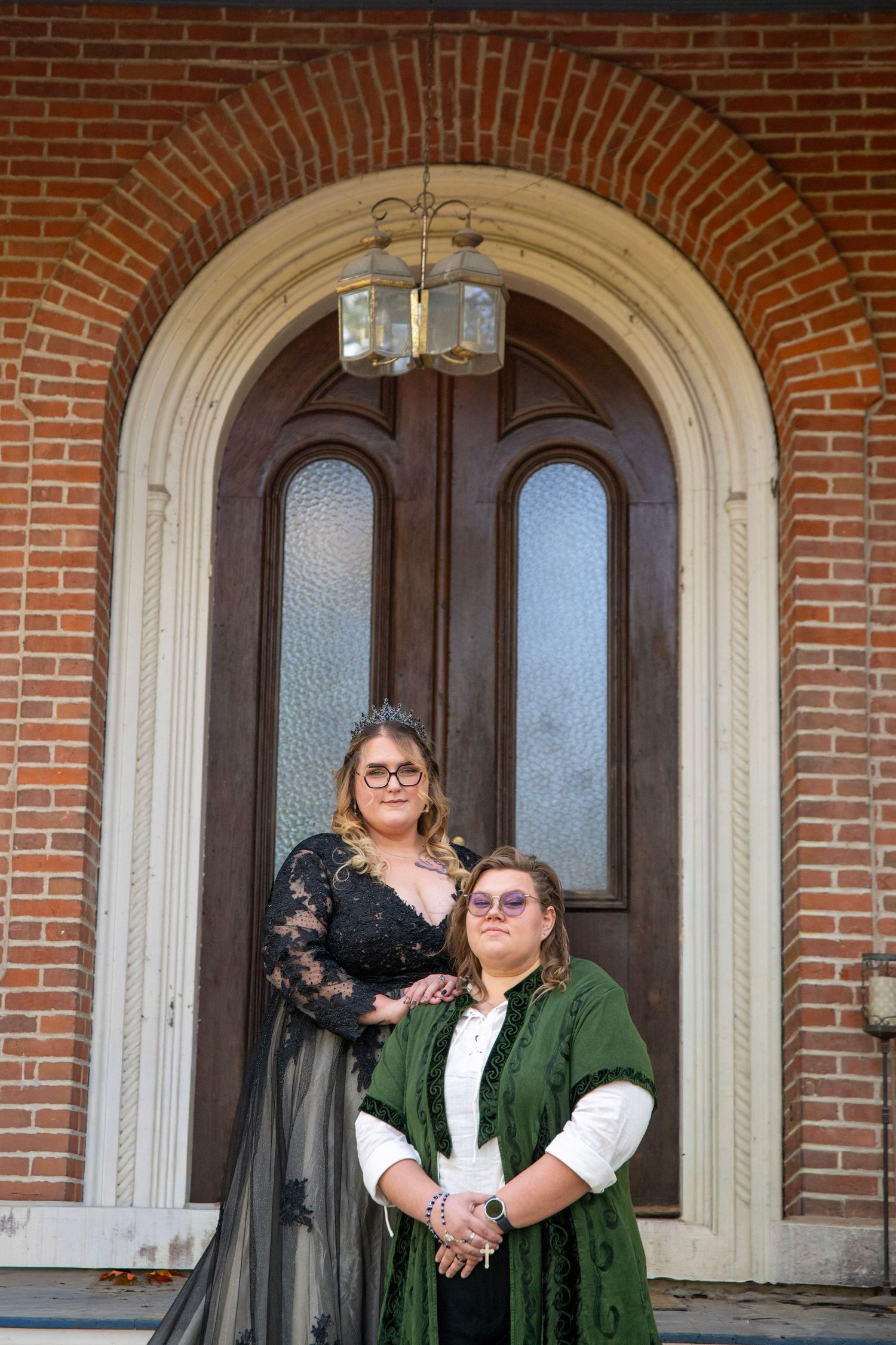 Two people in green and black outfits pose playfully on a brick building's front porch with arched doorways.