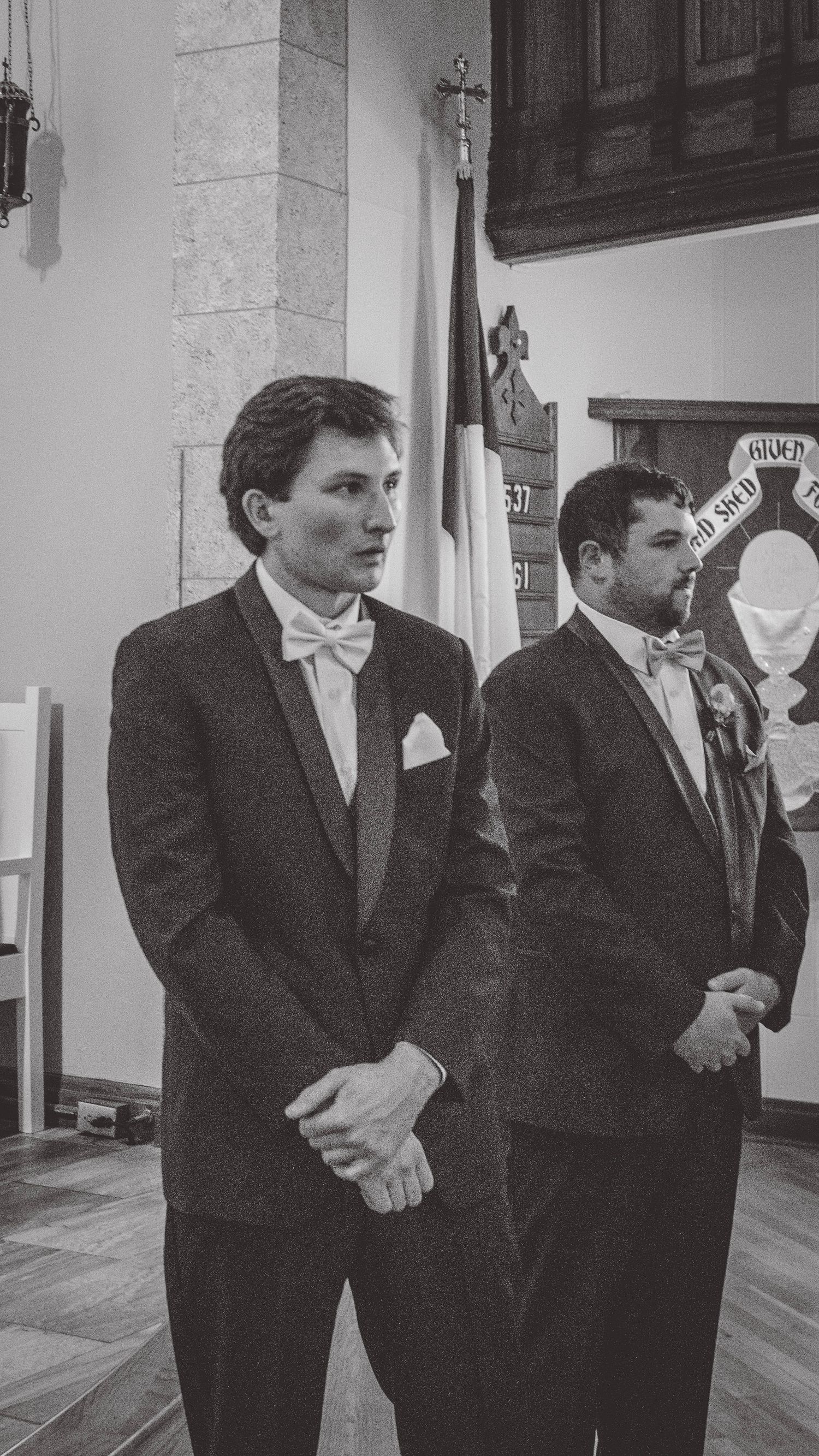 Groomsmen in black suits standing in a line at a formal event.