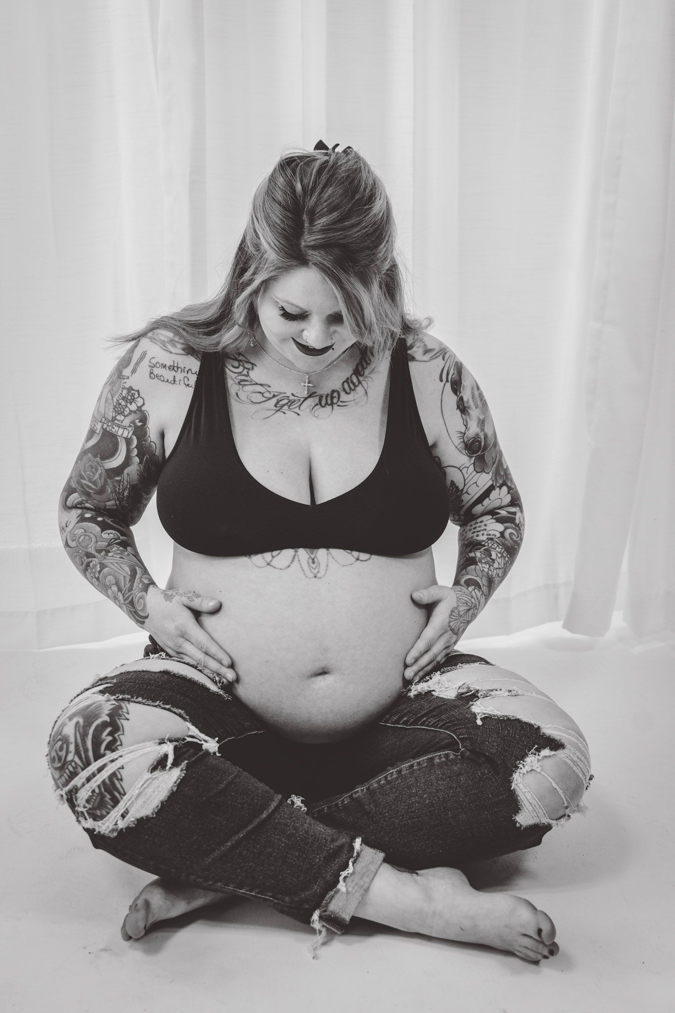 Artistic black and white maternity photo showing tattooed arms and crossed legs against white backdrop.