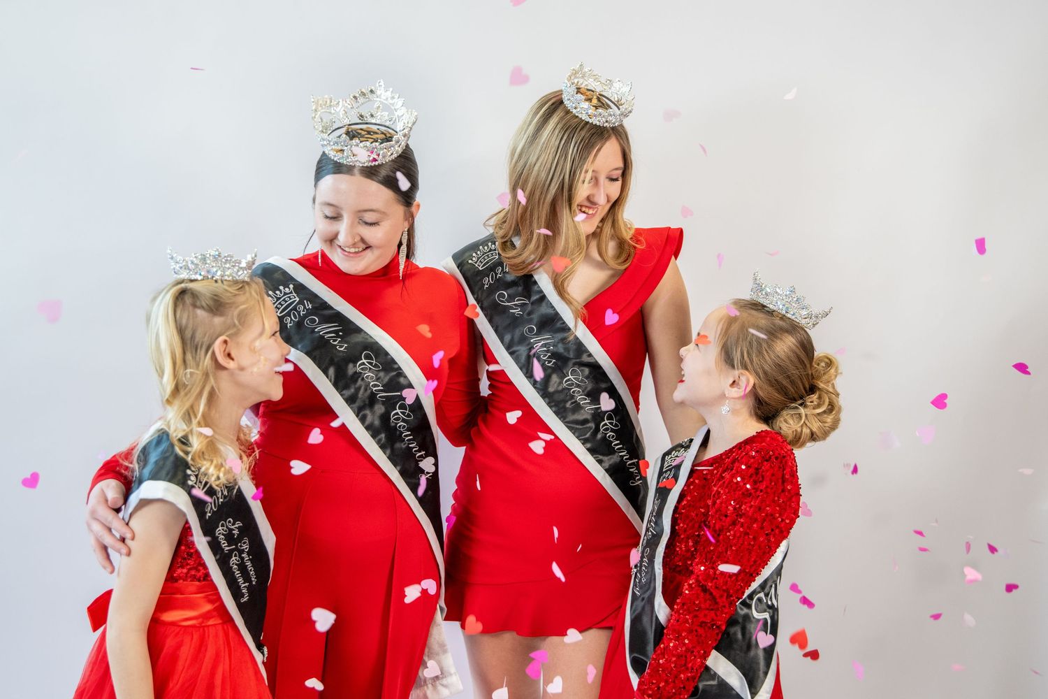 Group wearing red dresses and crowns pose against white backdrop with pink confetti.