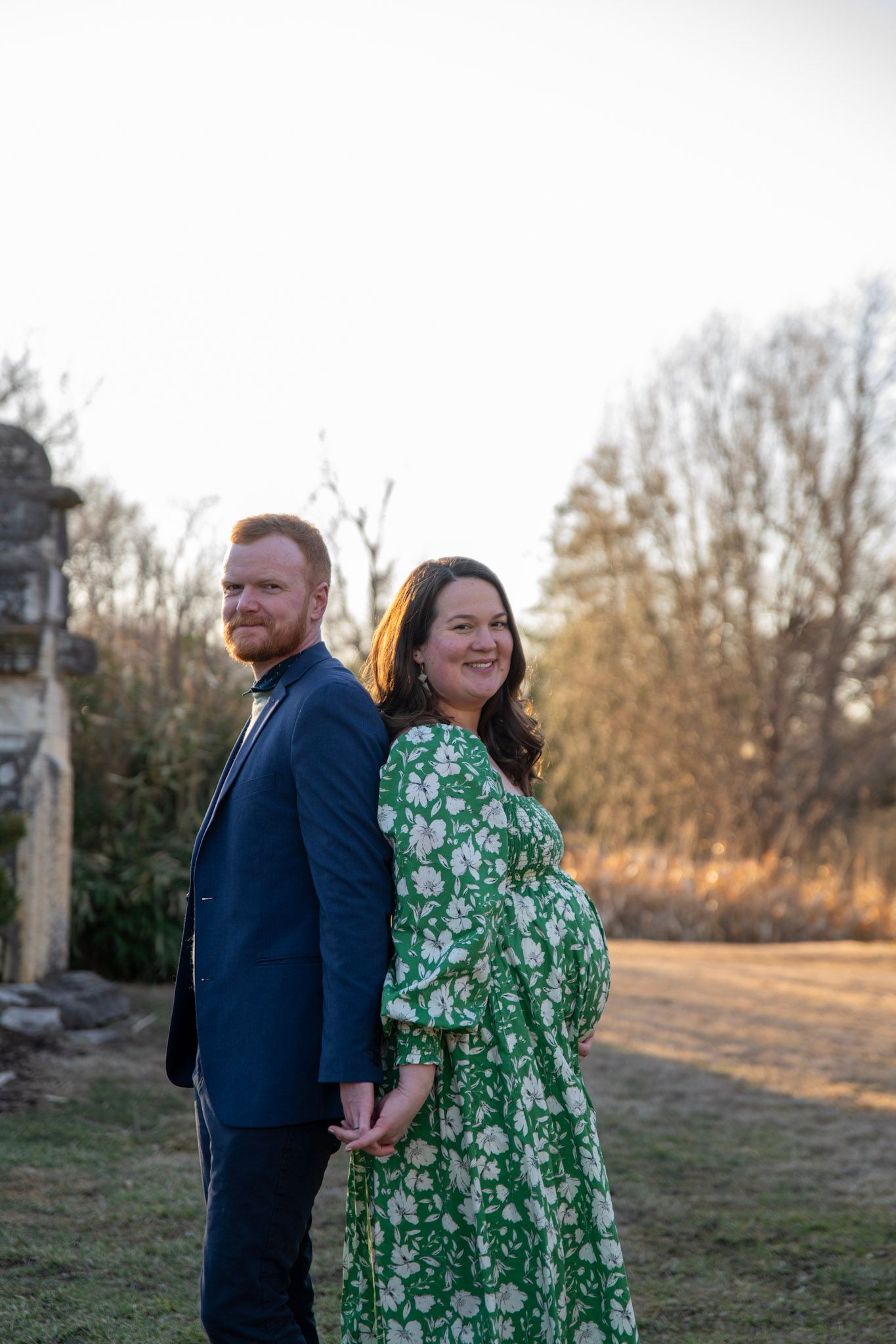 A couple poses outdoors at sunset, with the person in a green floral dress and navy suit embracing.