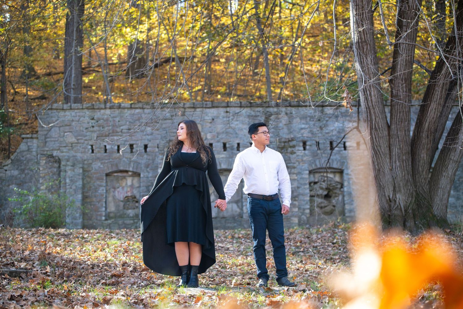 Couple poses near stone wall with golden sunlight filtering through trees.