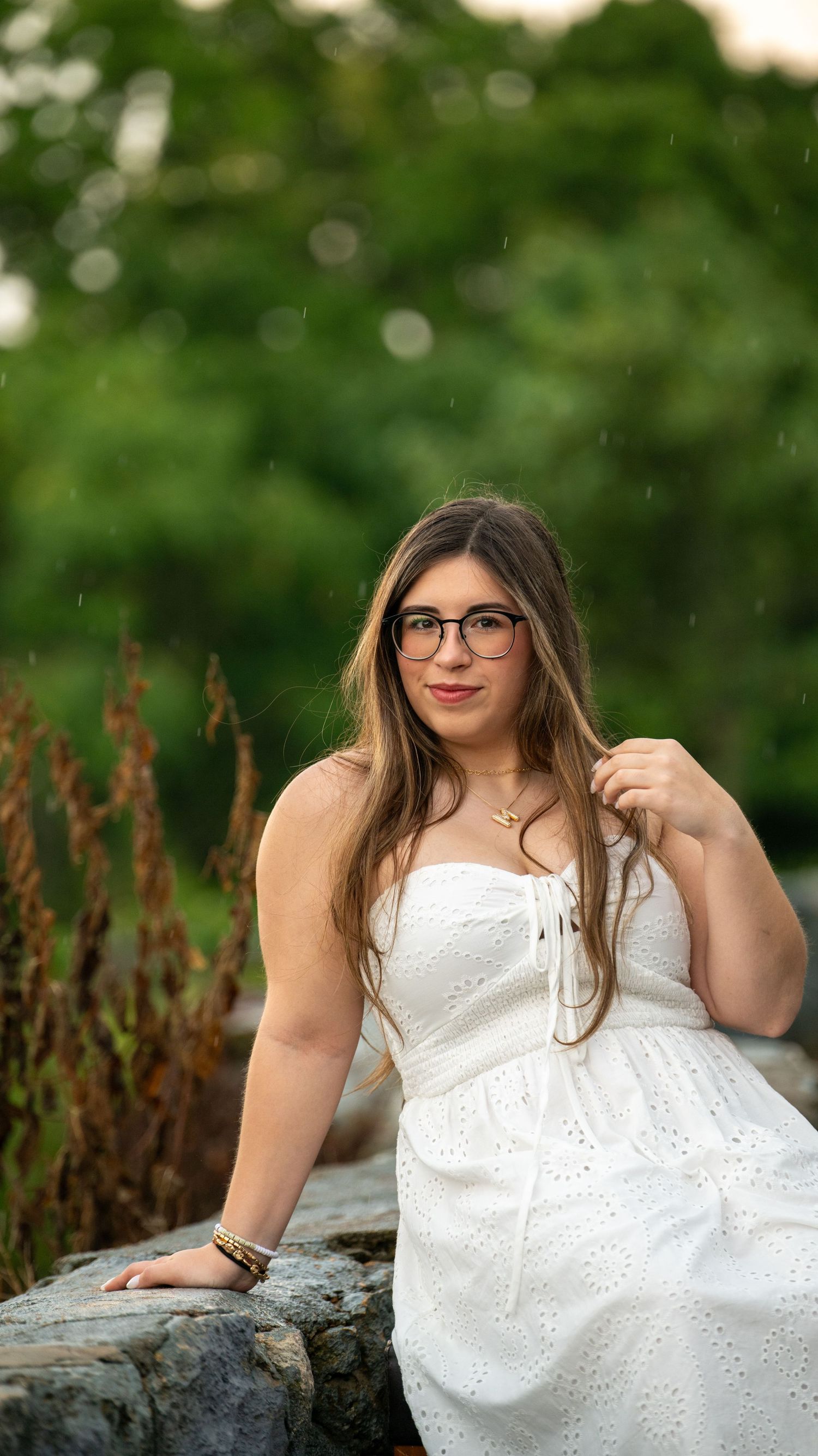 Person in white eyelet dress poses on stone wall with green foliage background.