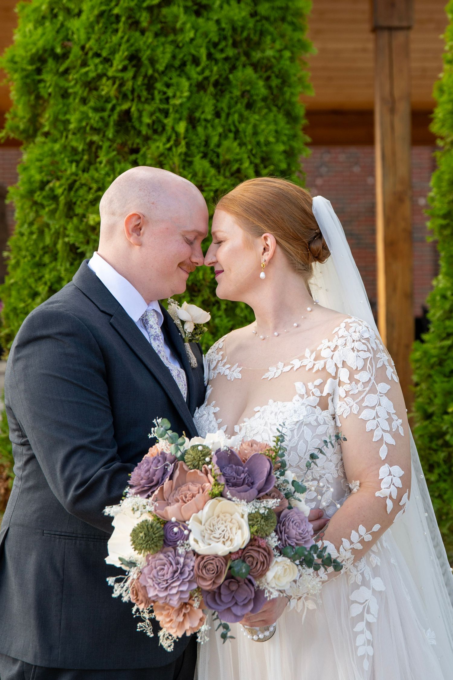 Wedding couple shares intimate moment with purple and cream bouquet against green foliage backdrop.