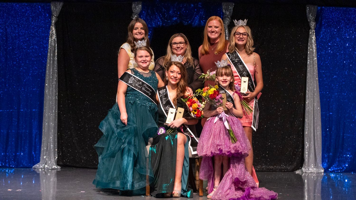 Pageant contestants in colorful gowns stand together during an awards ceremony.