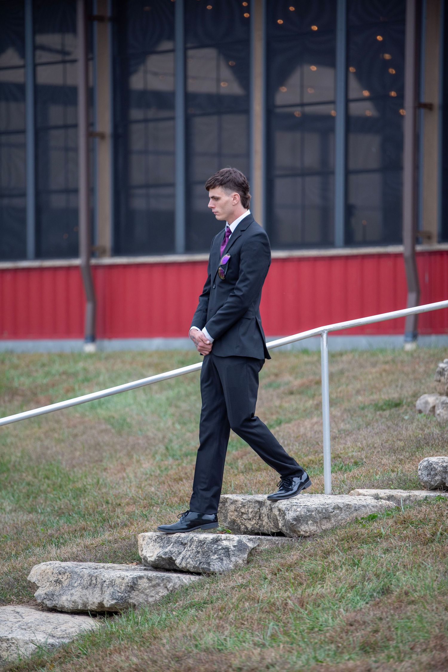 Person in formal black suit walking along an outdoor stone wall with railing.