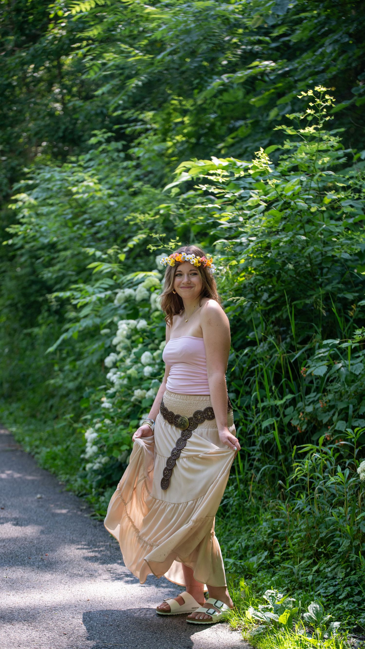 A person in a pink dress and flower crown poses playfully on a forest path.