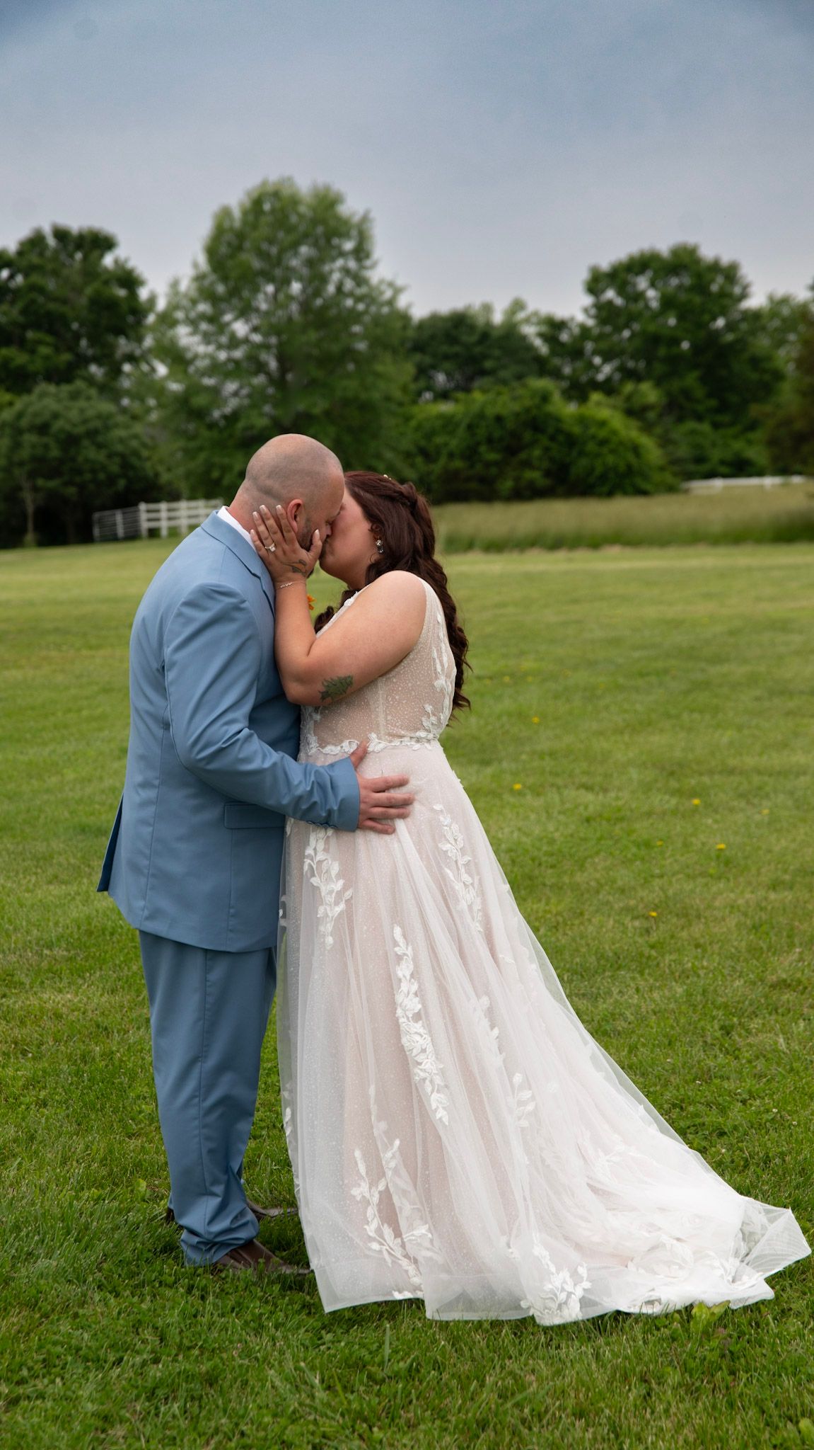 A romantic wedding moment captured on a green lawn with a light blue suit and white lace dress.