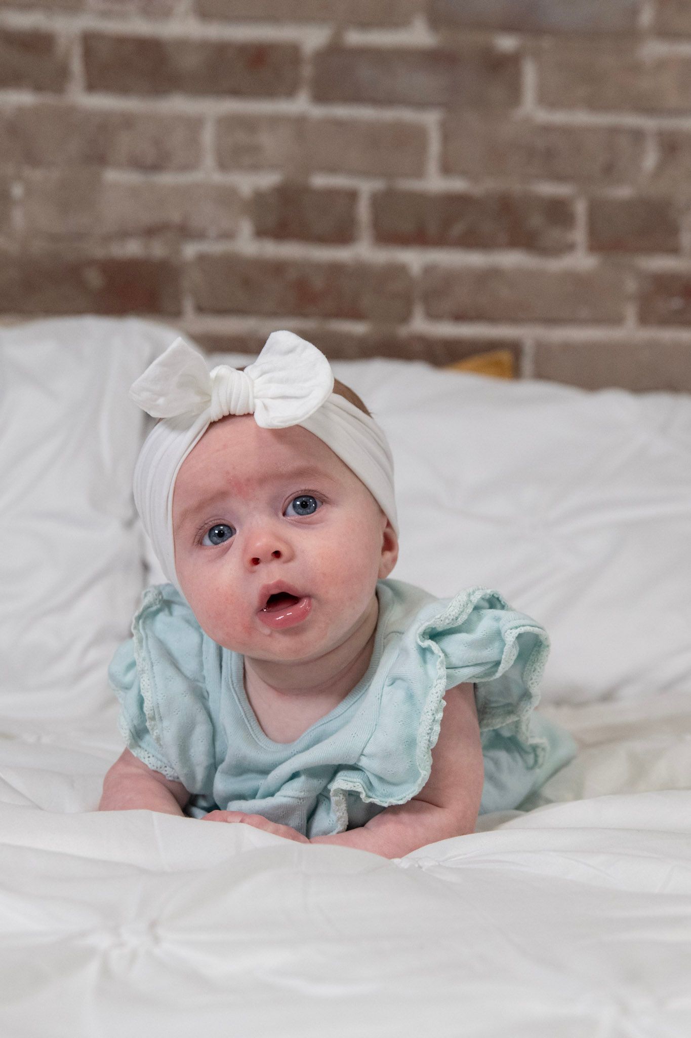 Sweet baby in mint green ruffled dress and white bow headband lying on white bedding against brick wall.