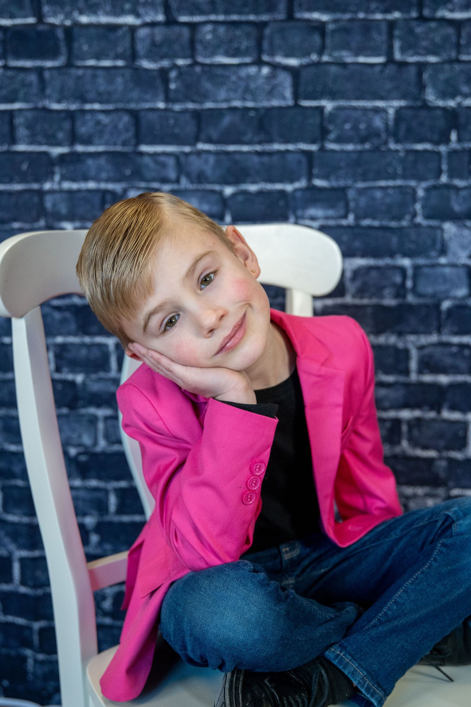 Casual pose on a white chair wearing a vibrant pink jacket and jeans against brick wall.