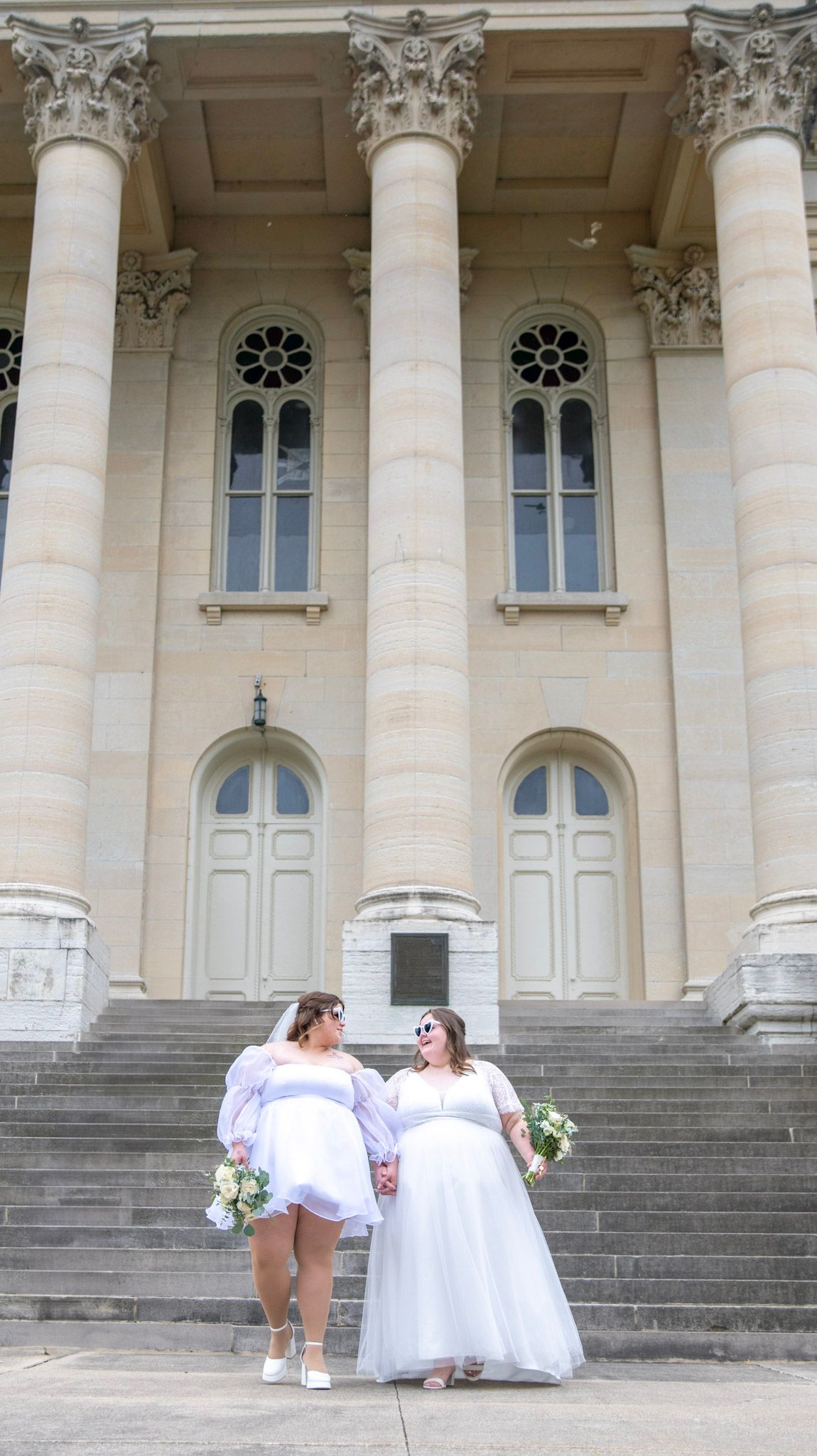 A couple in white dresses shares a moment on the steps of a grand columned building.