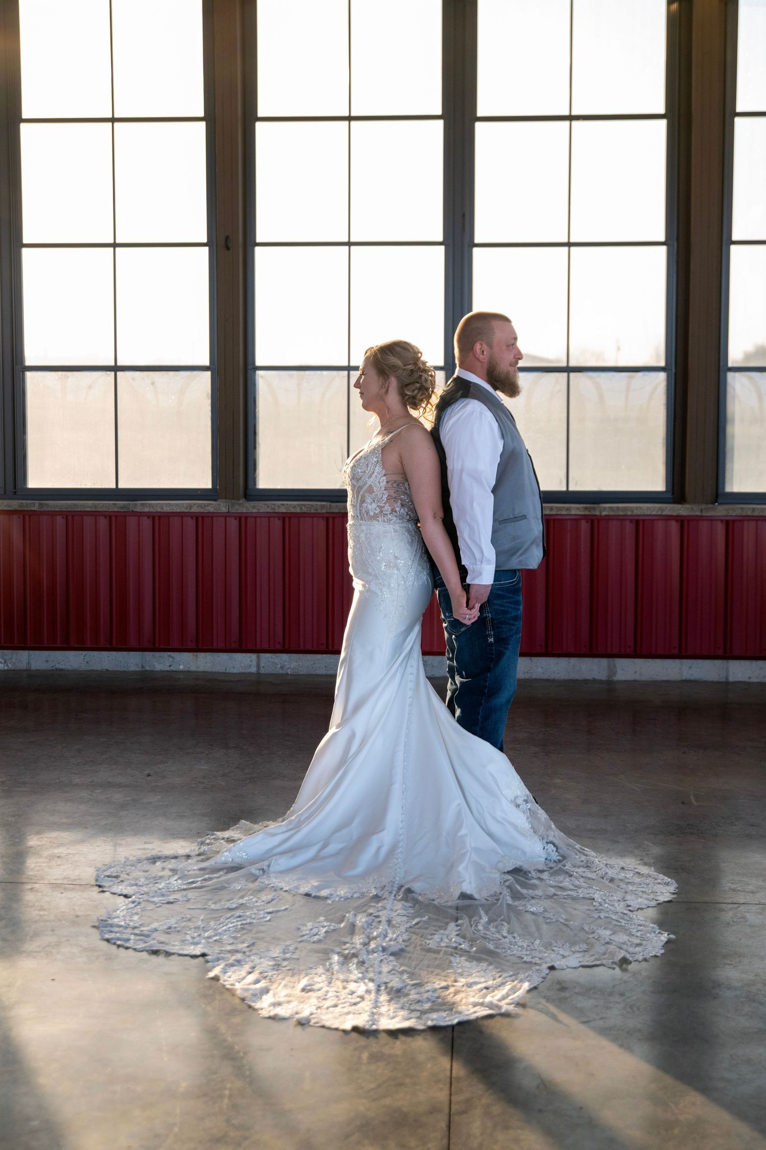A wedding couple stands back-to-back in formal attire near large windows.