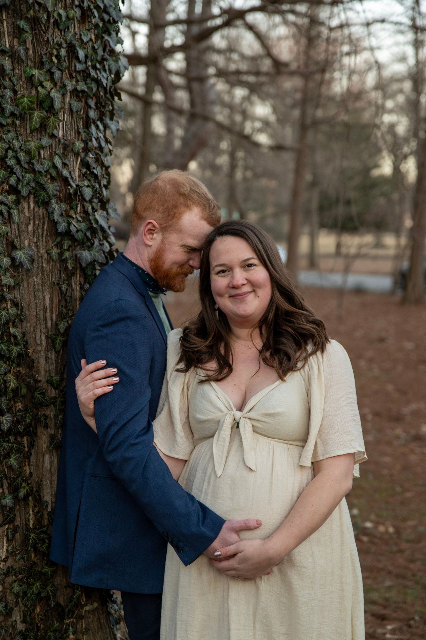 A couple embraces by a tree during a maternity photoshoot in an outdoor setting.