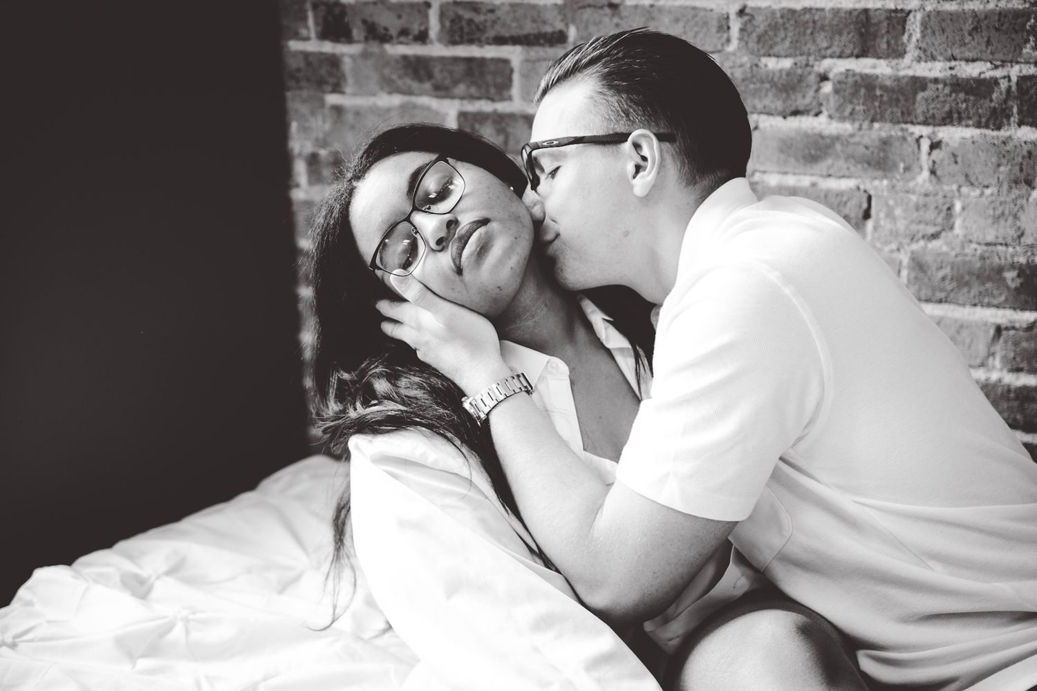 Intimate moments captured in monochrome as couple lies on white bed against brick wall.