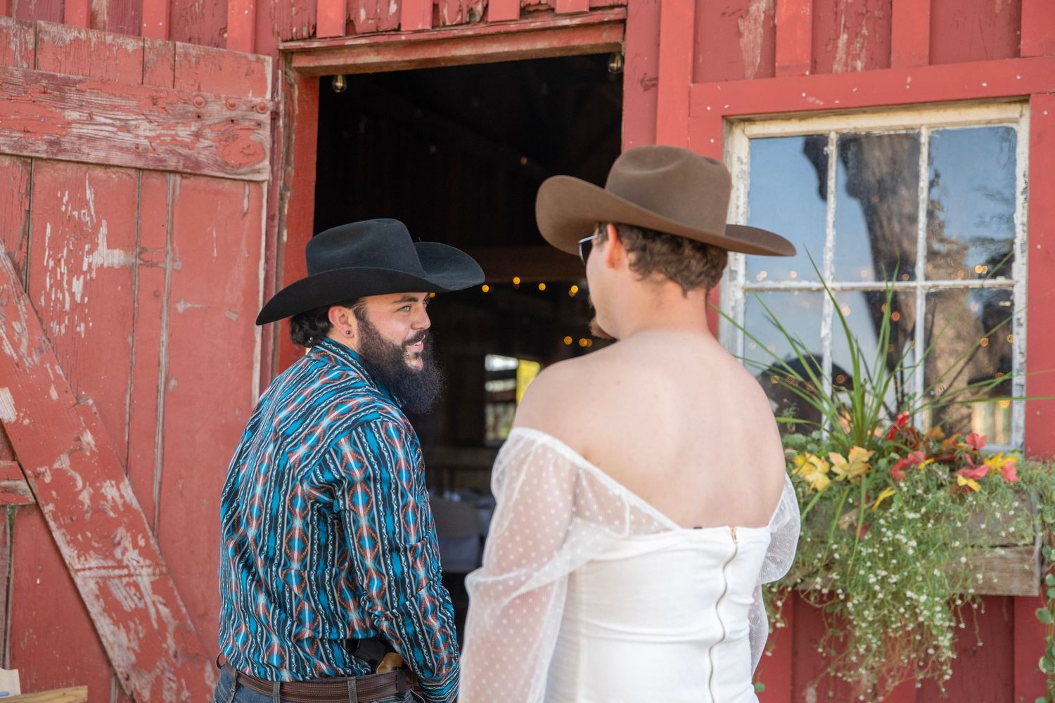 Couple poses for photos in front of a weathered red barn with string lights overhead.