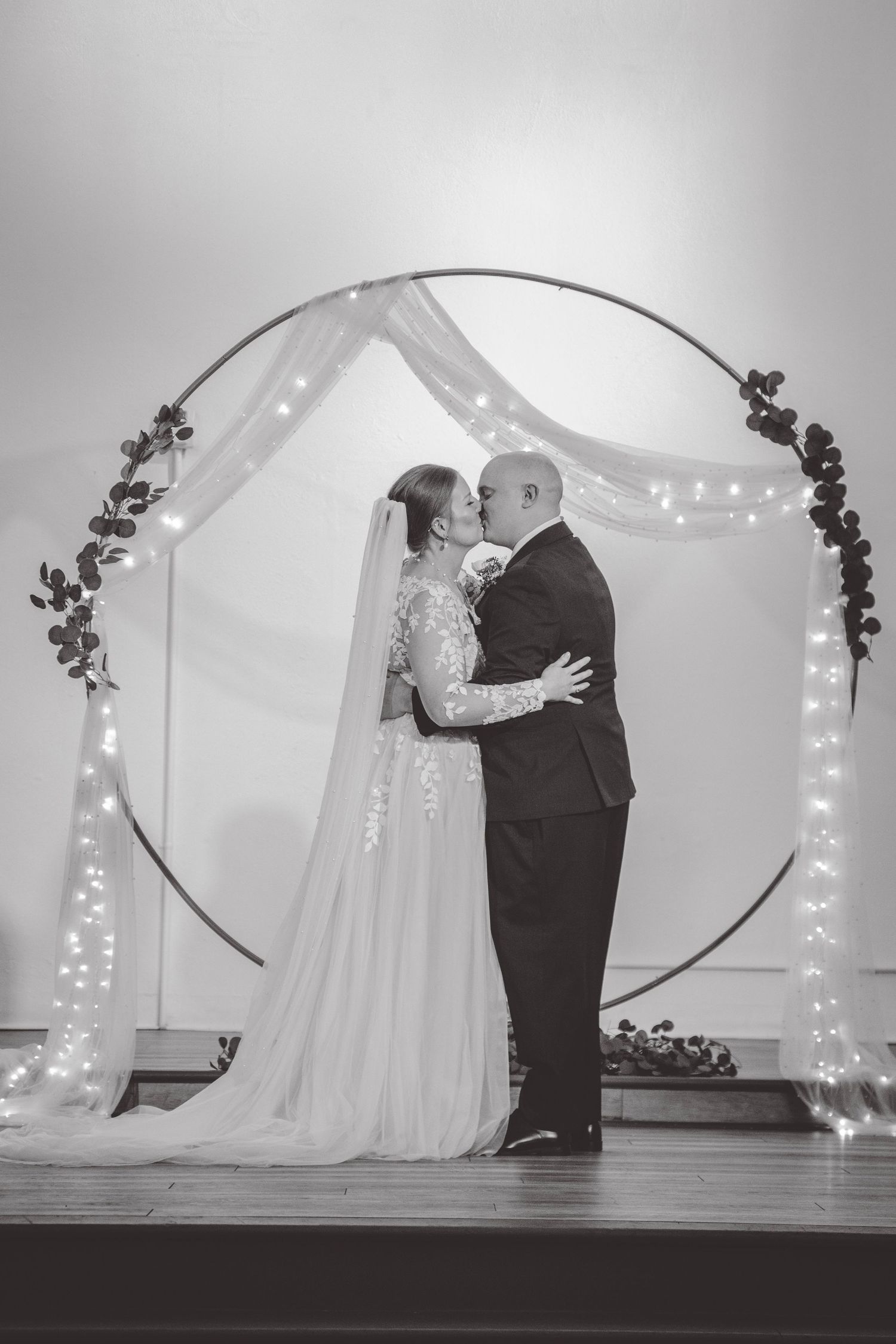Wedding couple stands under illuminated circular arch decorated with greenery and fairy lights.