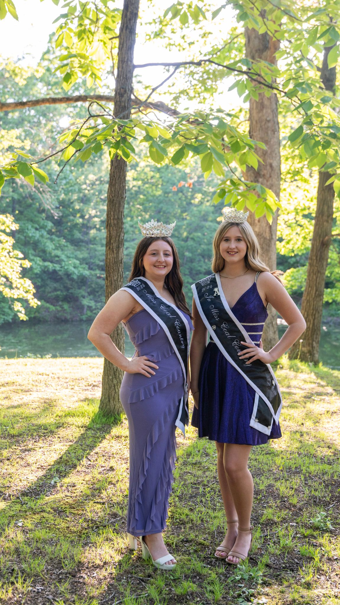 Two pageant contestants in purple and navy dresses pose together in a sunlit wooded area.