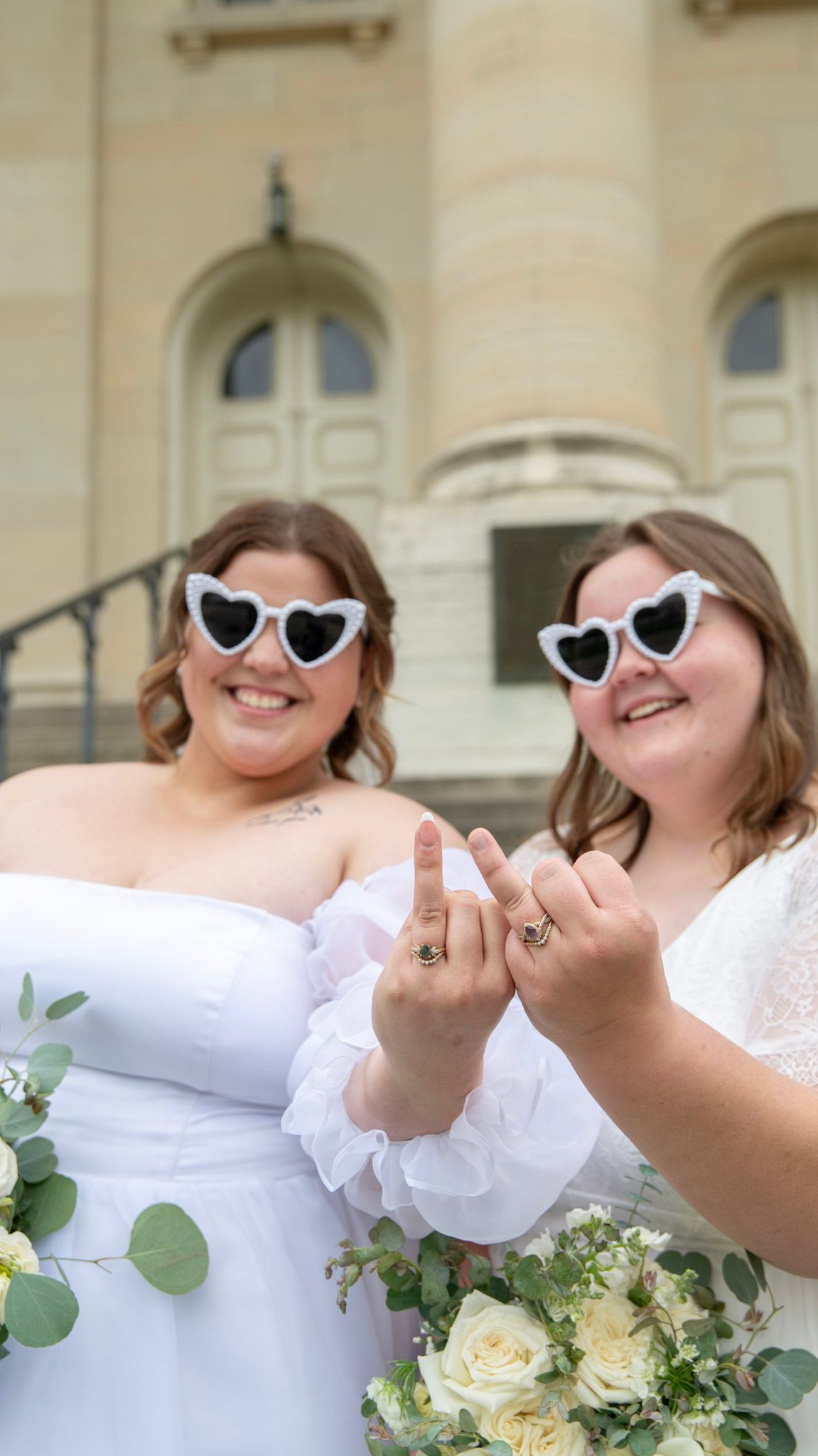 Playful wedding couple wearing heart sunglasses share laughs with white bouquets.