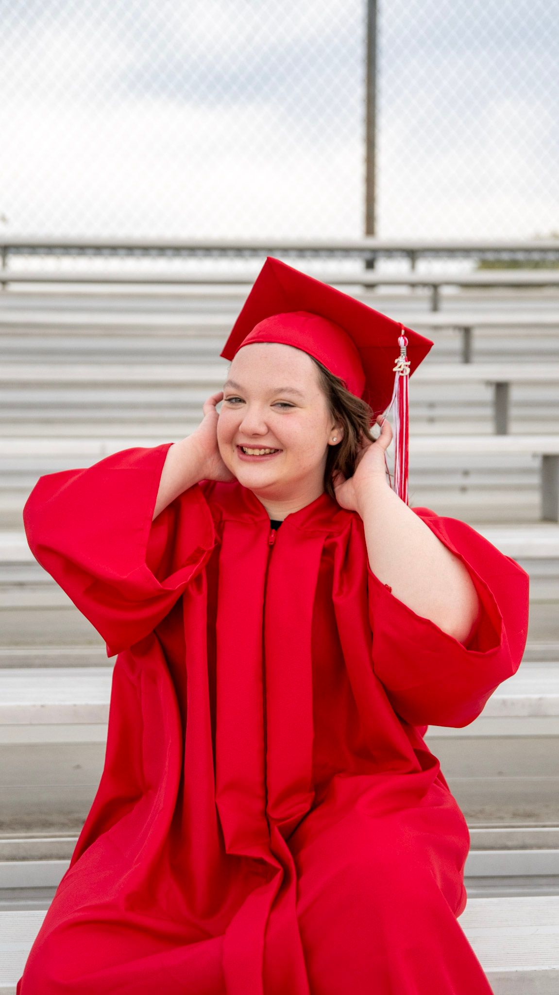 Graduate in red graduation regalia poses joyfully on metal bleachers.
