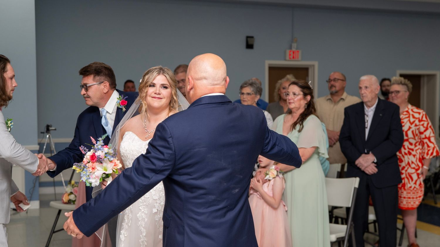 A wedding ceremony moment in a reception hall with guests seated in white chairs.