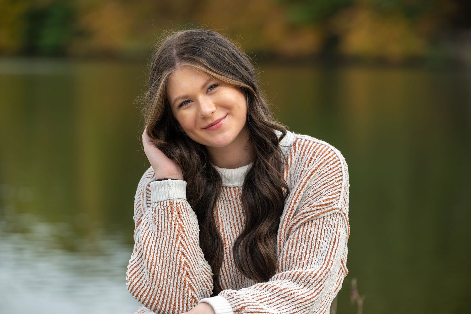 Person in beige sweater smiling warmly by waterfront during fall season.
