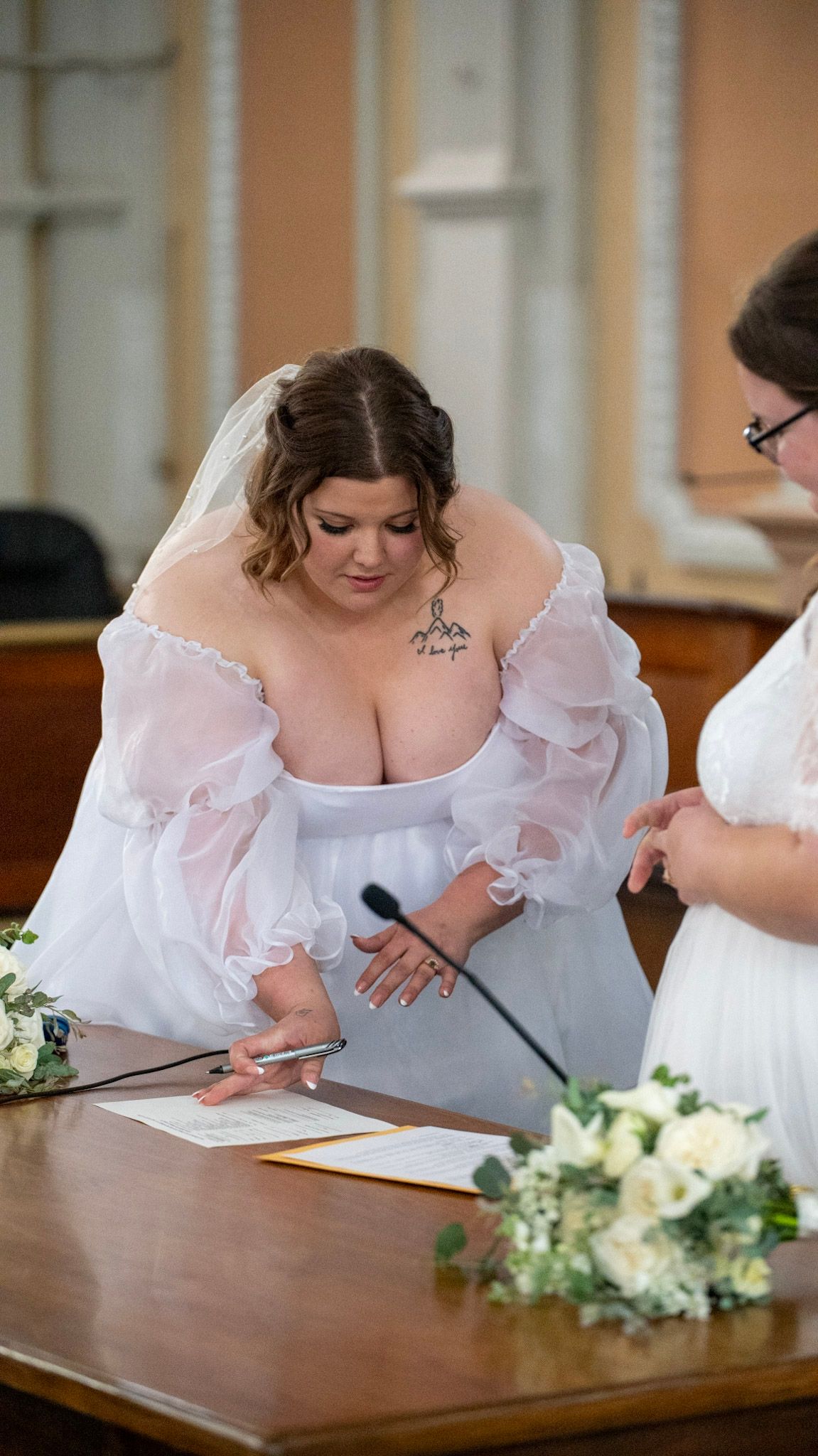 Person in white dress signing marriage documents at wooden table with floral bouquet.