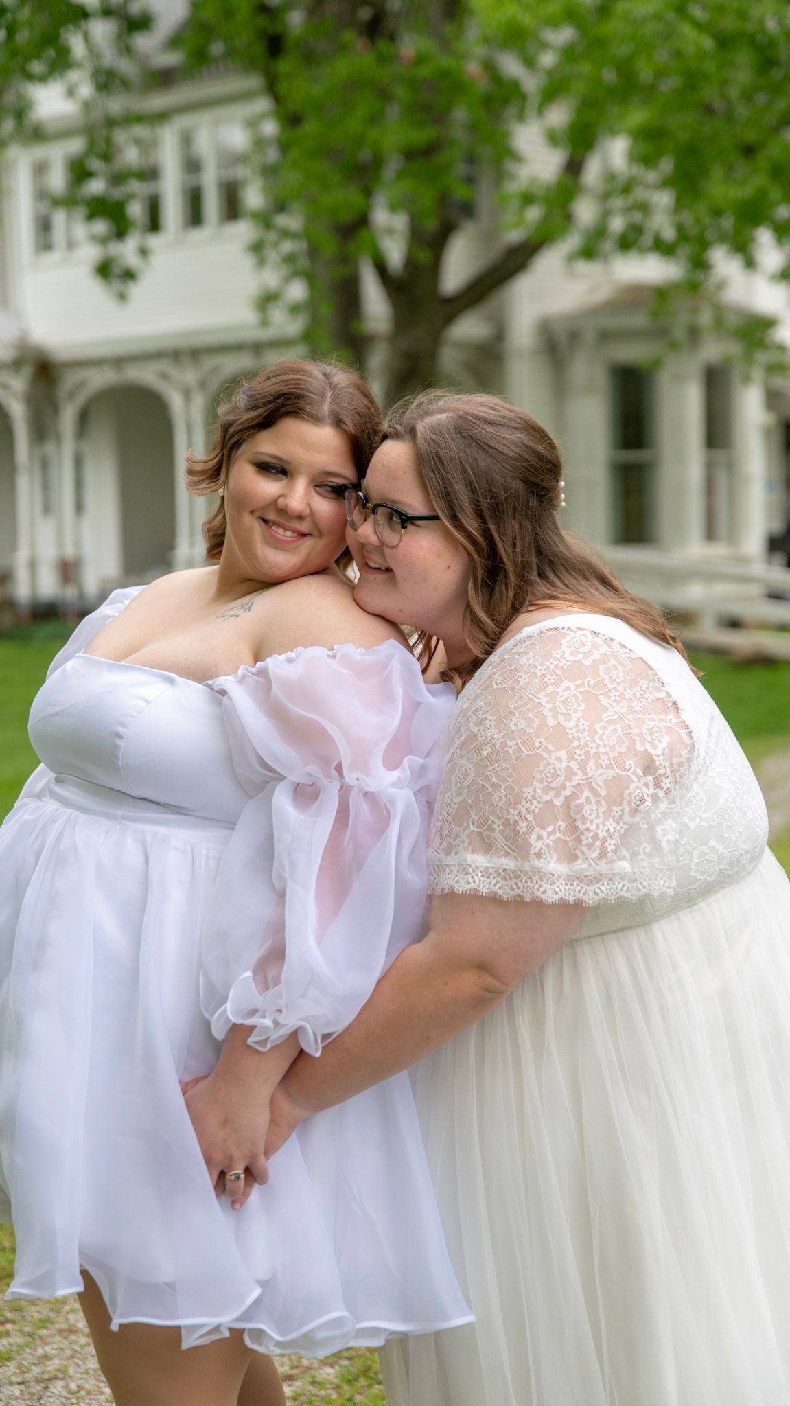 Two people in white dresses share an intimate moment outdoors in front of a classic white building.