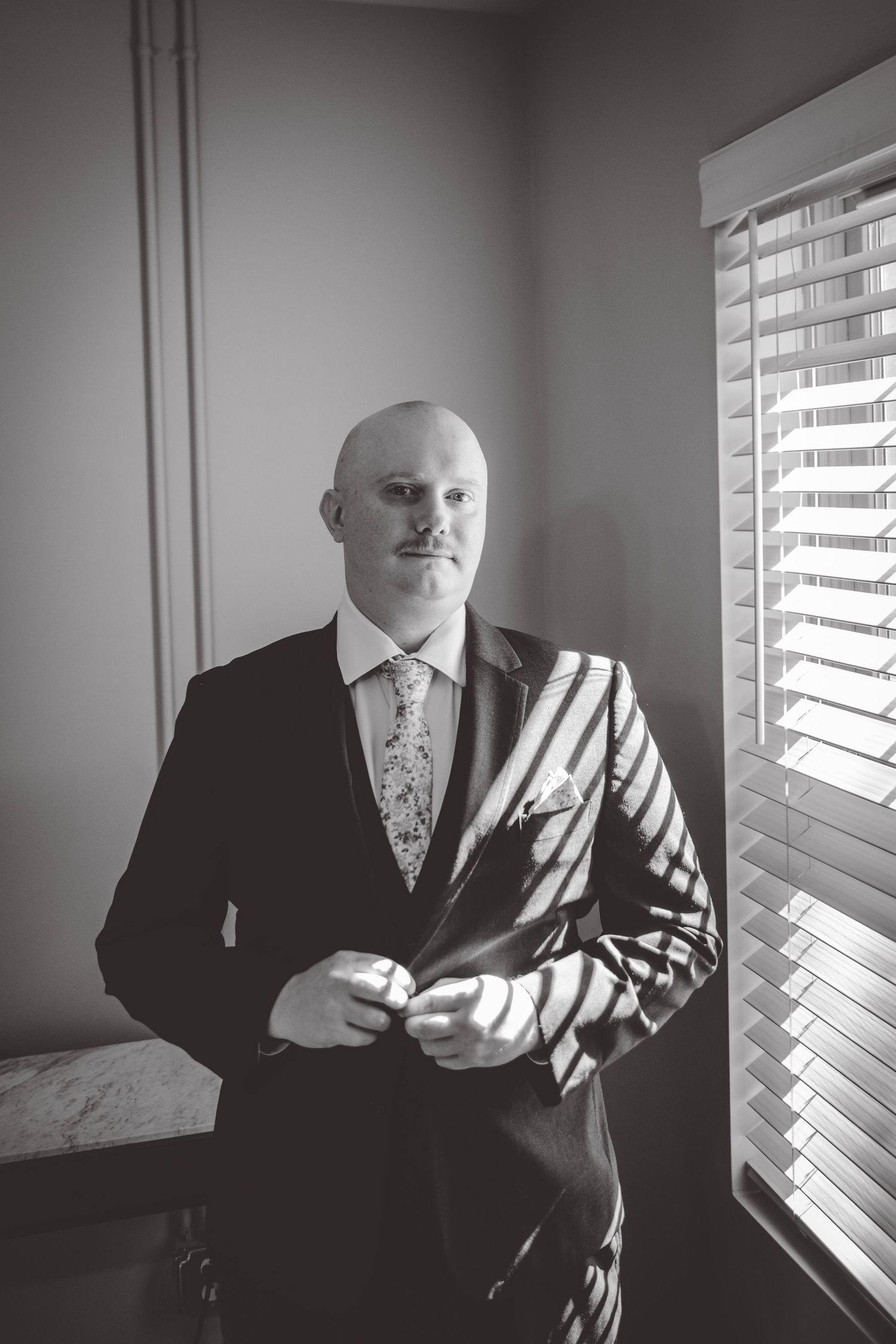 Dramatic black and white portrait by window with venetian blinds casting film noir style shadows.