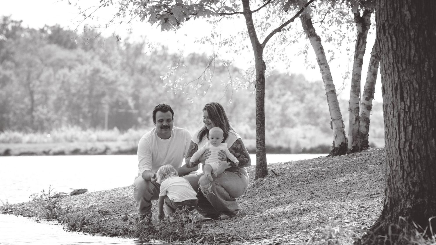 Black and white panoramic view of people relaxing by trees near water.