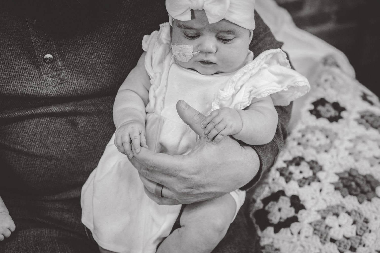 Black and white photo of baby in white dress being held against patterned quilt.