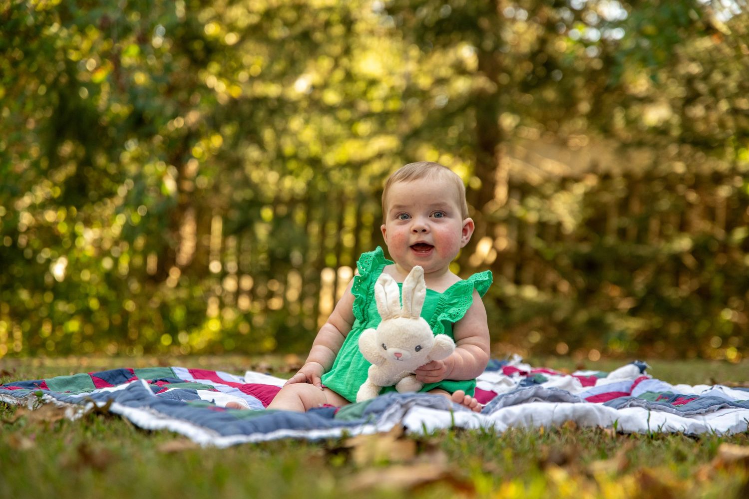 Smiling baby in green outfit holding stuffed bunny on patterned blanket outdoors.