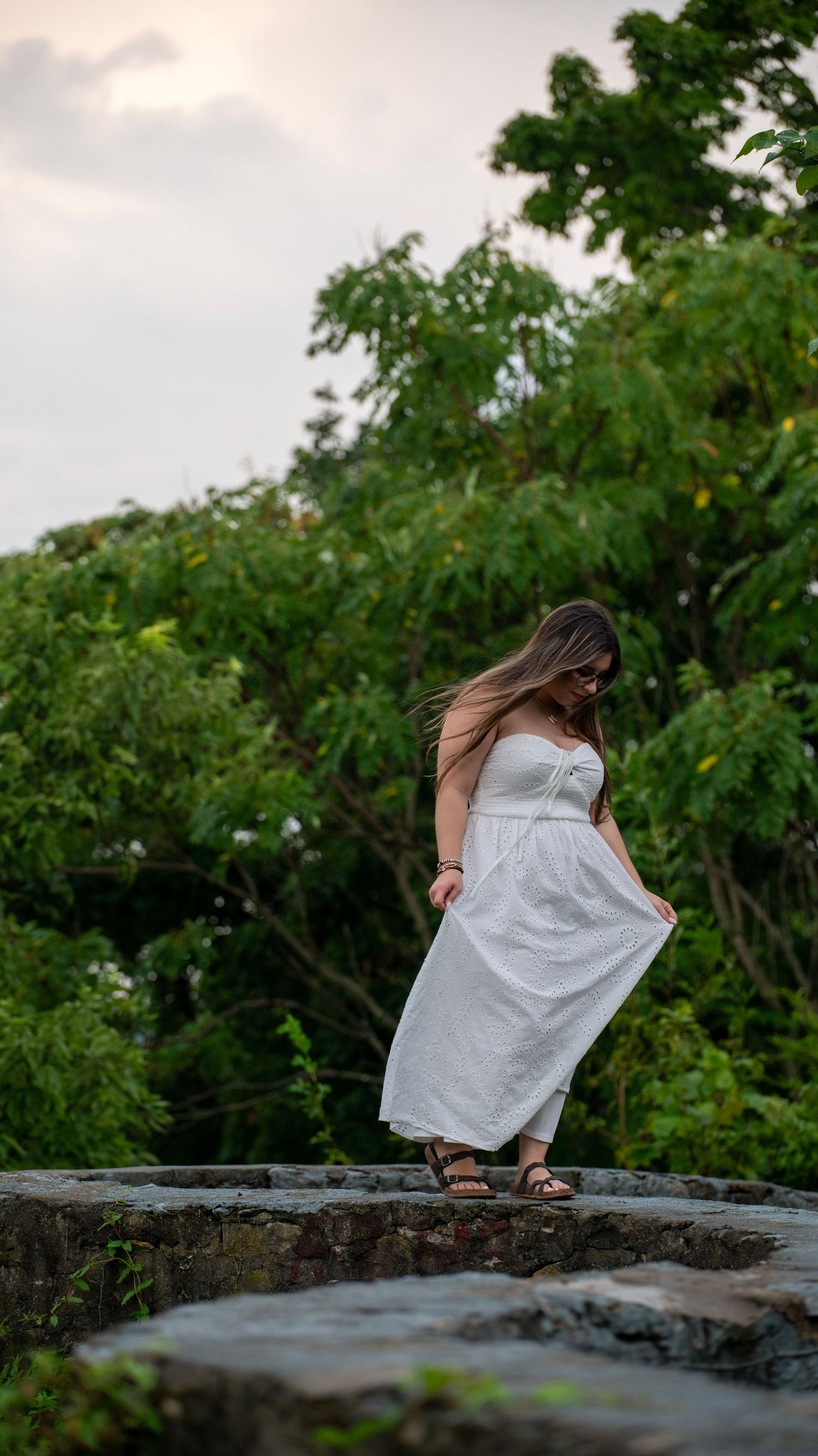 A figure in a flowing white dress stands gracefully on stone steps surrounded by lush greenery.