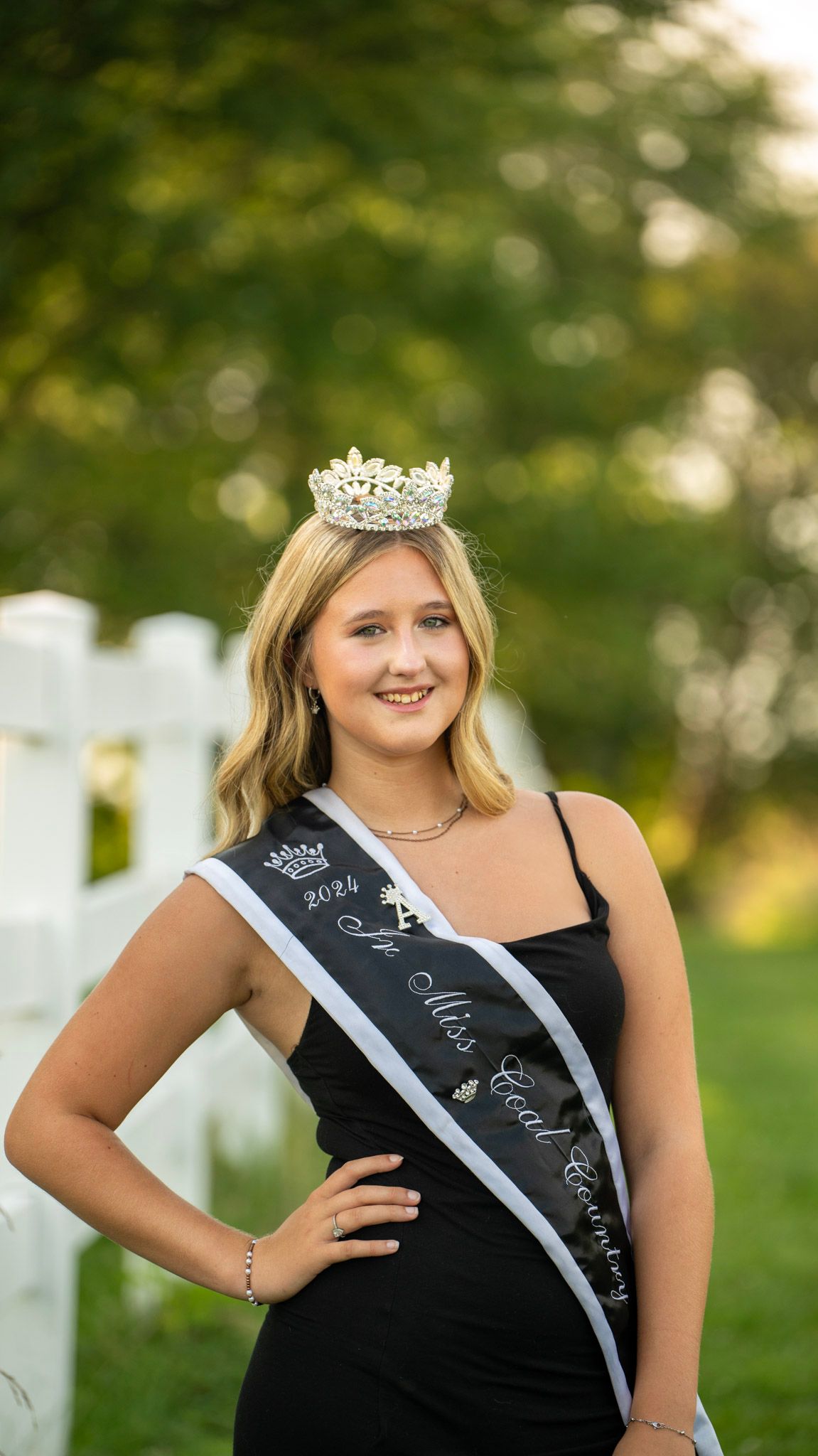 A crowned pageant contestant in a black dress smiles while posing by a white fence.
