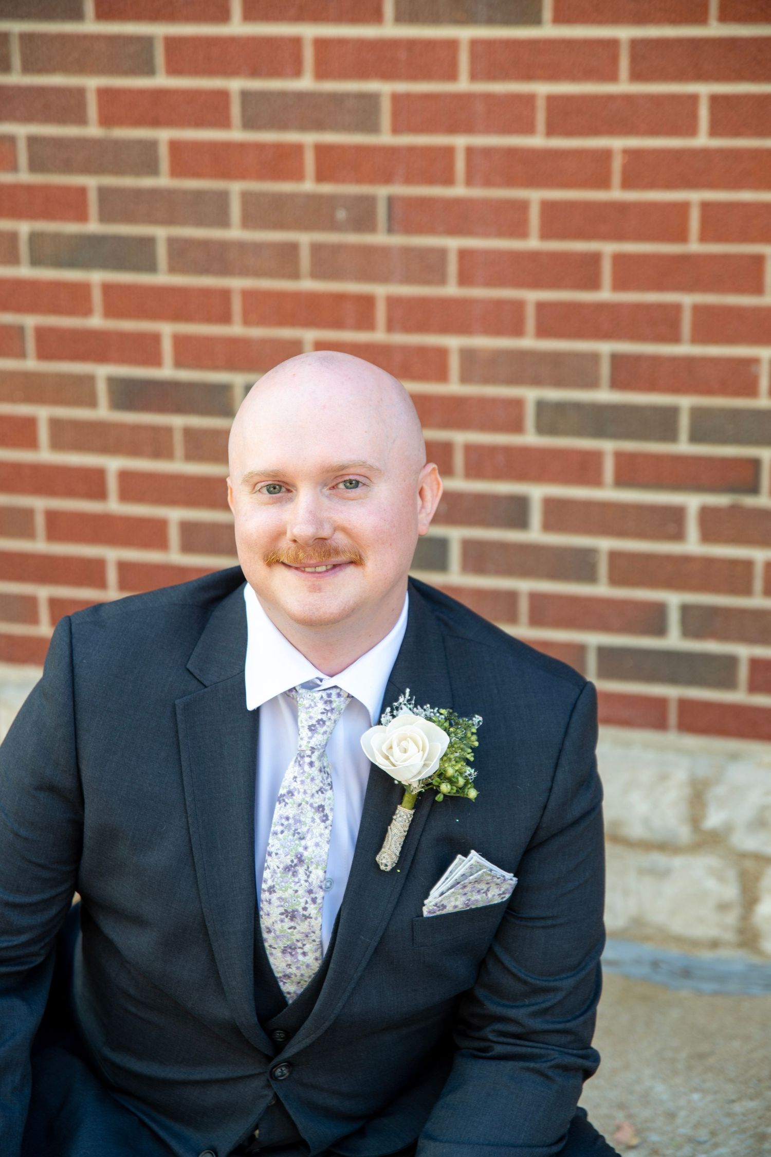 Portrait series of a groom in a dark suit with floral tie and white boutonniere against brick background.