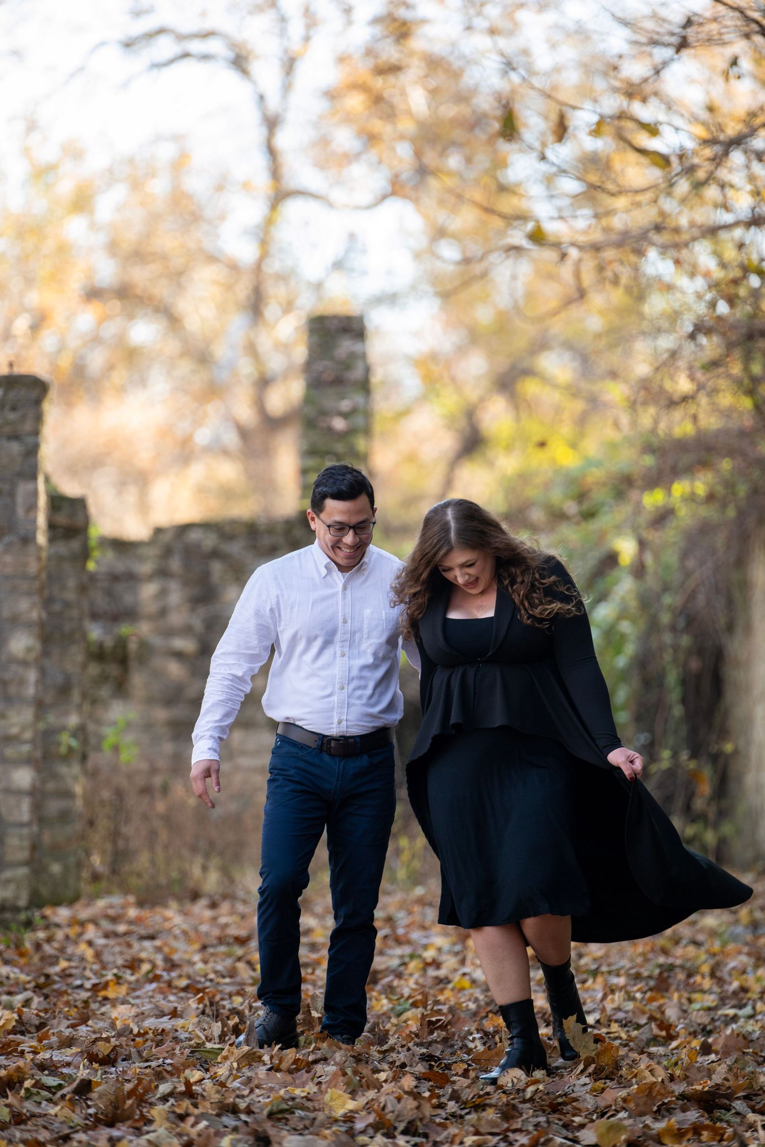 Couple walks playfully through fallen leaves near historic stone wall.