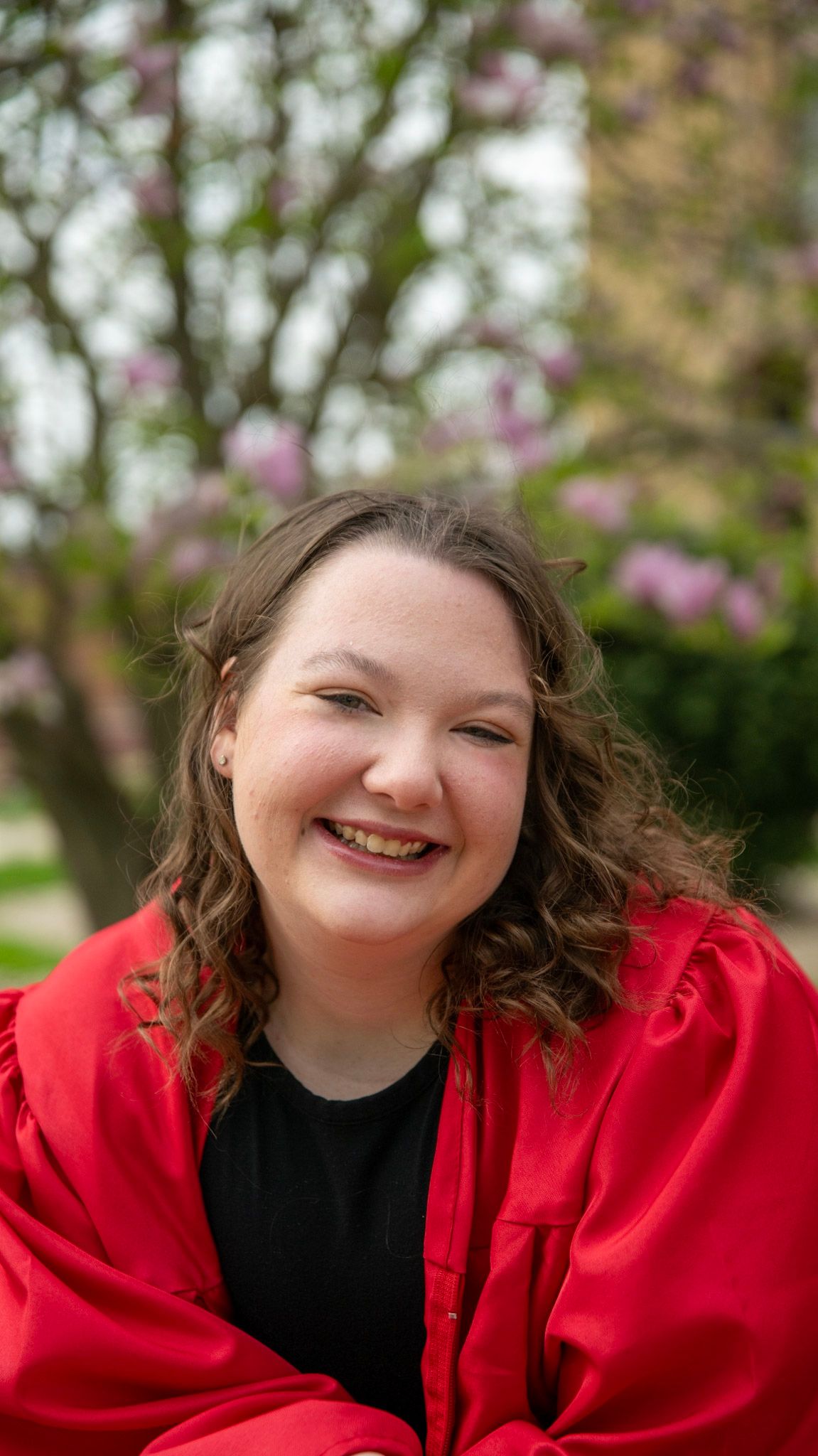 Graduate in red gown smiles joyfully against blooming spring trees.