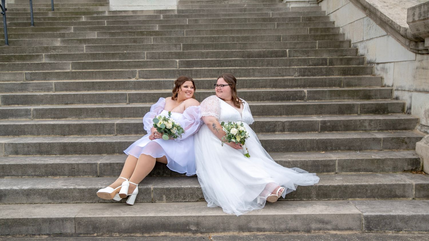 Newlyweds in white dresses sitting together on grand stone steps with bouquets.