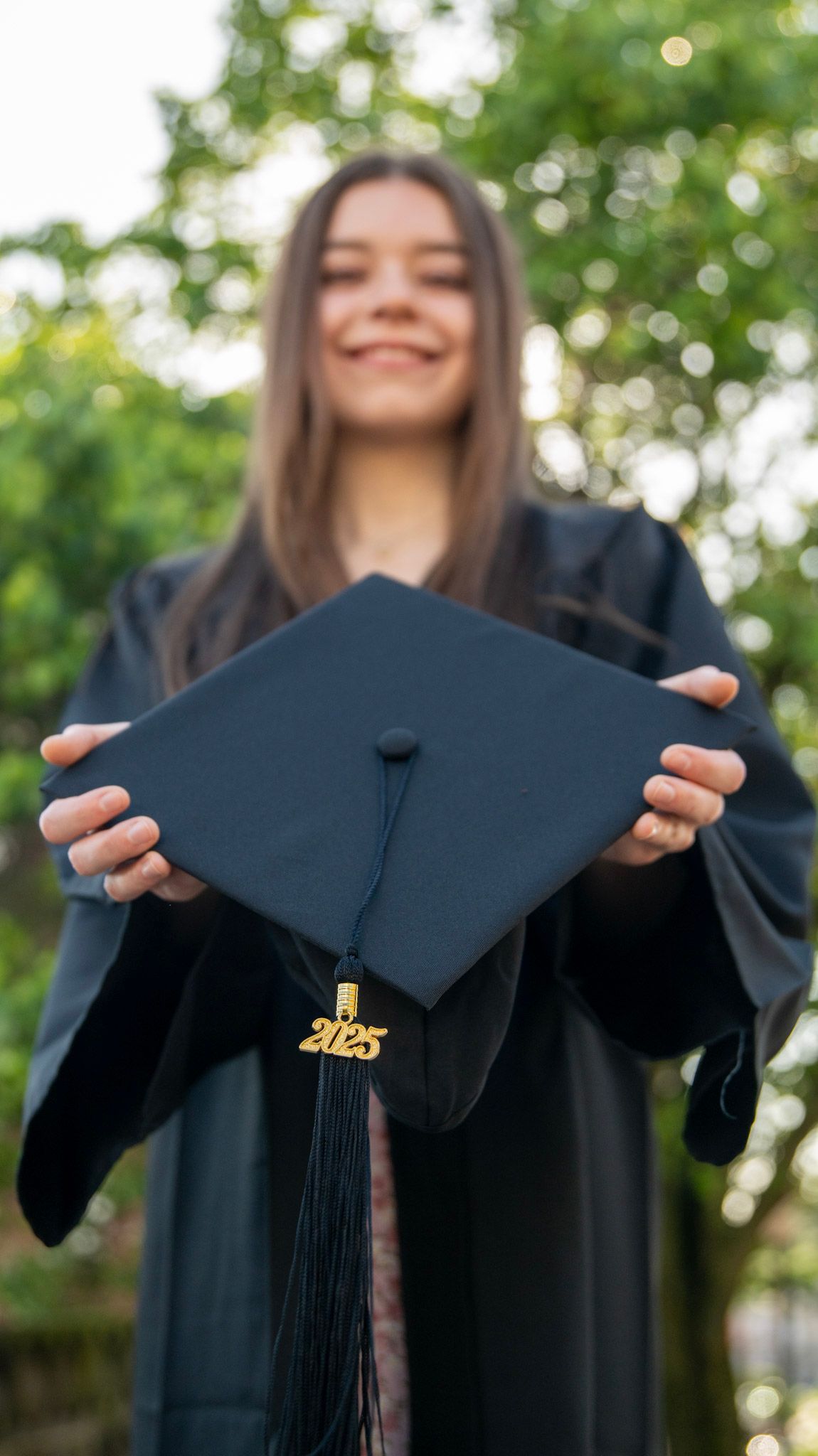 Graduate holding up a black graduation cap with 2023 tassel against green foliage.