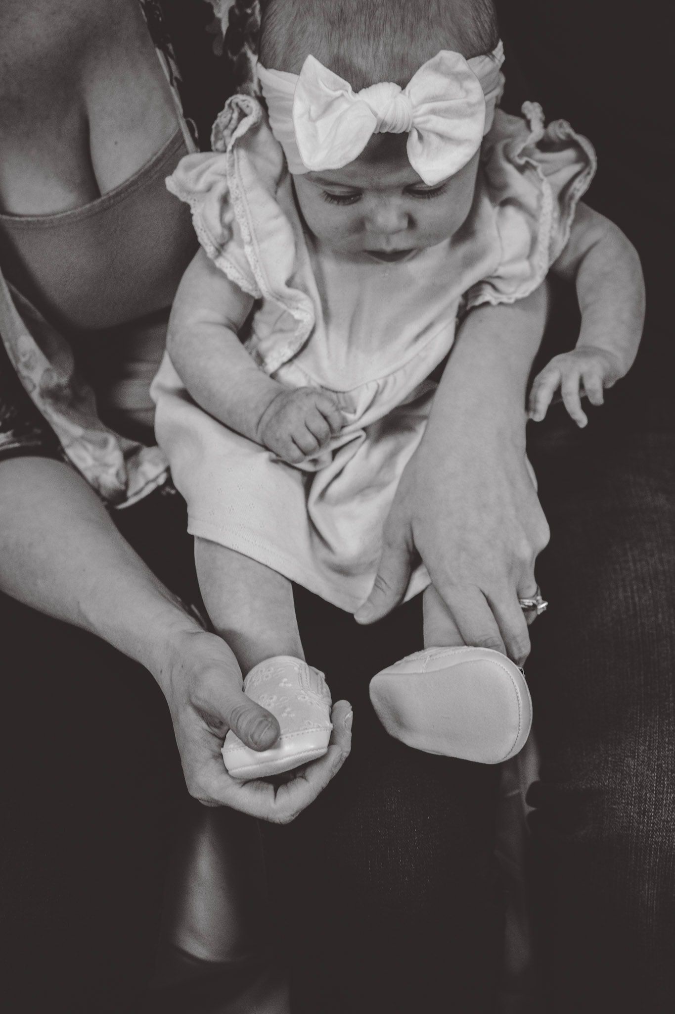 Black and white photo of baby in ruffled dress and bow being held on lap.