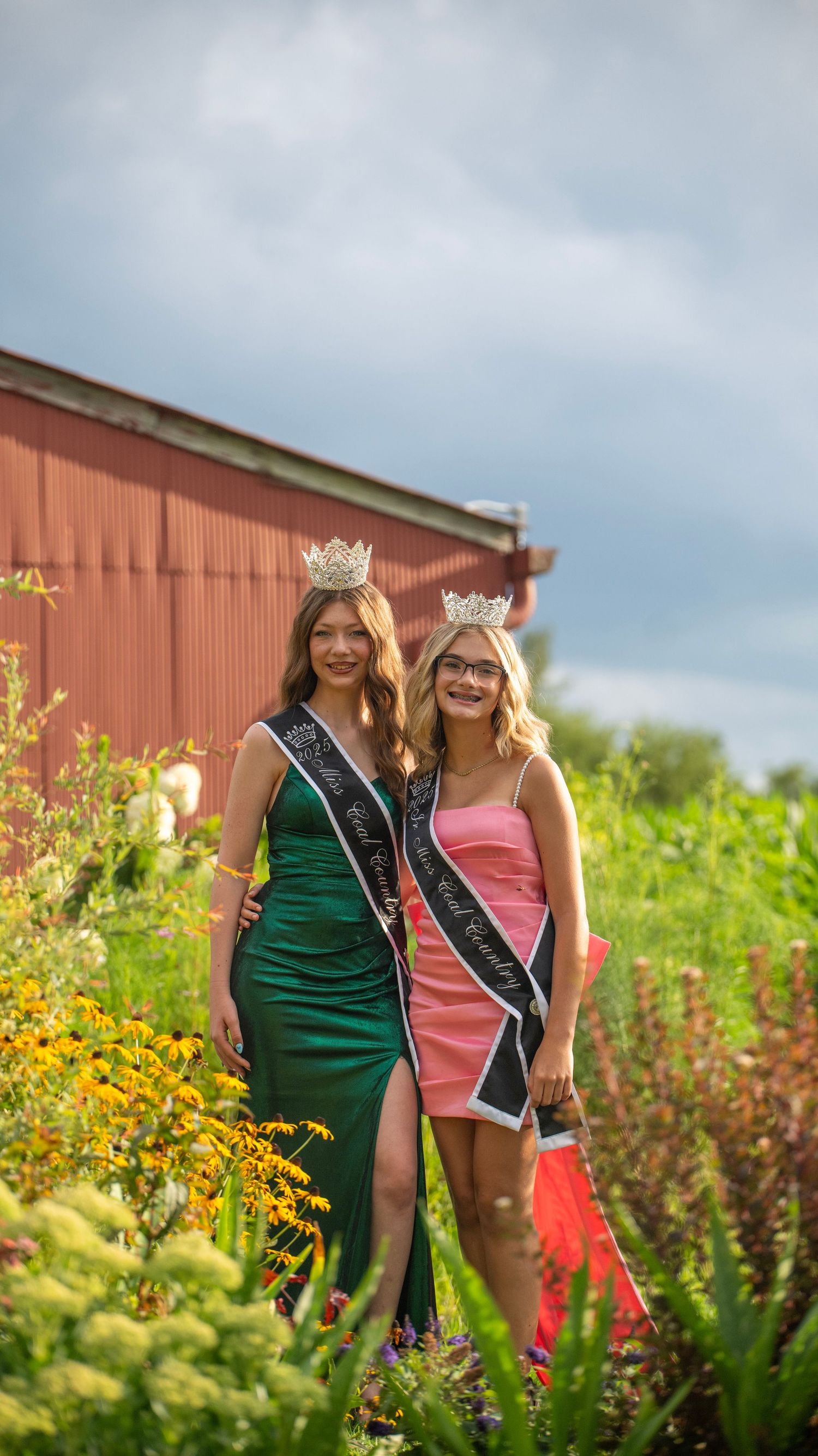 Two pageant winners in green and pink dresses pose together in flowering garden.