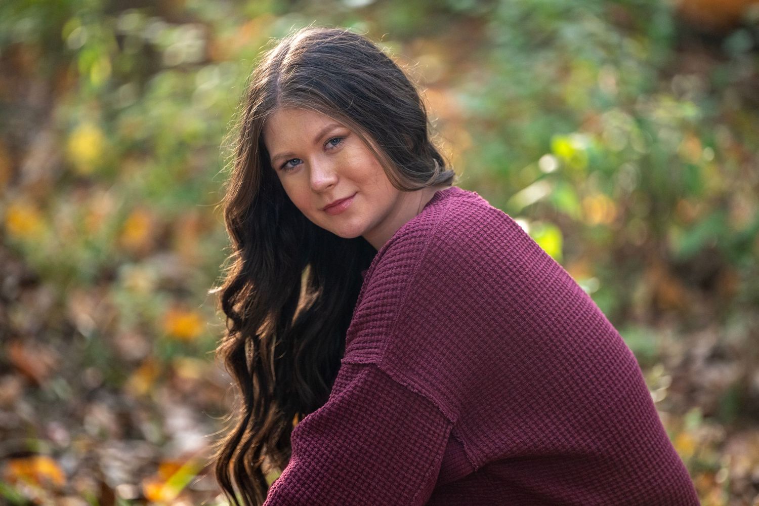 Person in maroon sweater poses against blurred fall foliage background.