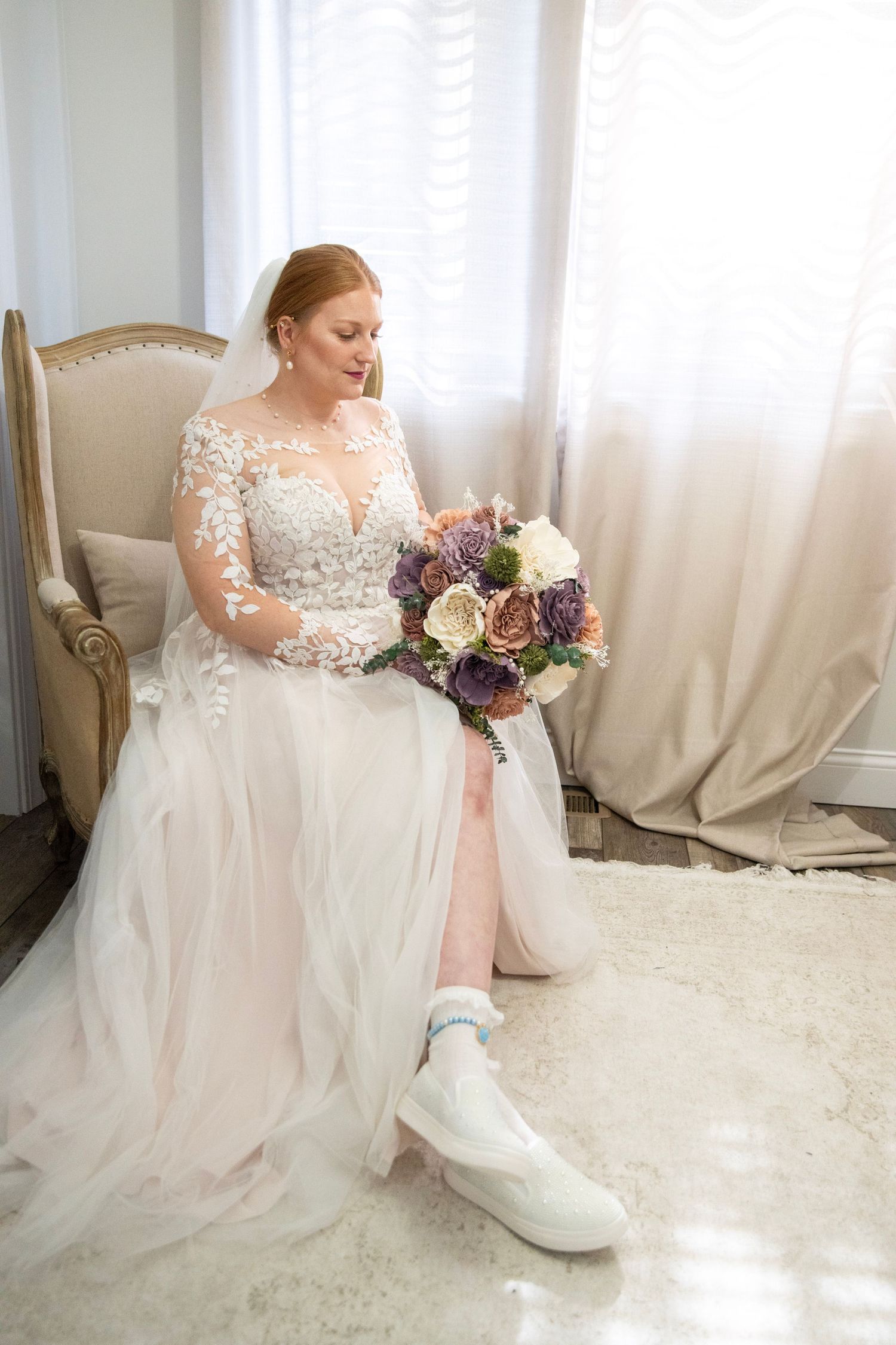 Bride in lace wedding gown sits elegantly with bouquet in bright white room.