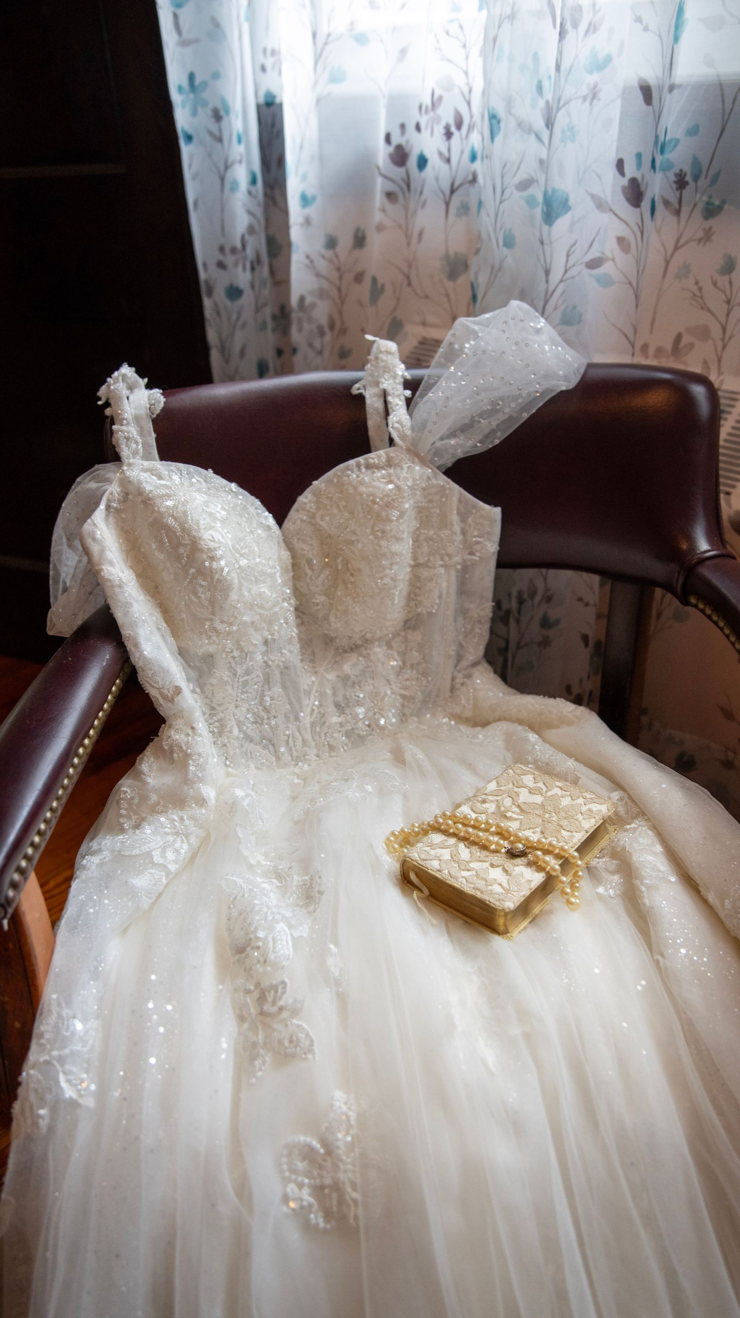 Detailed shot of an elegant white lace wedding dress with book accent on leather chair.